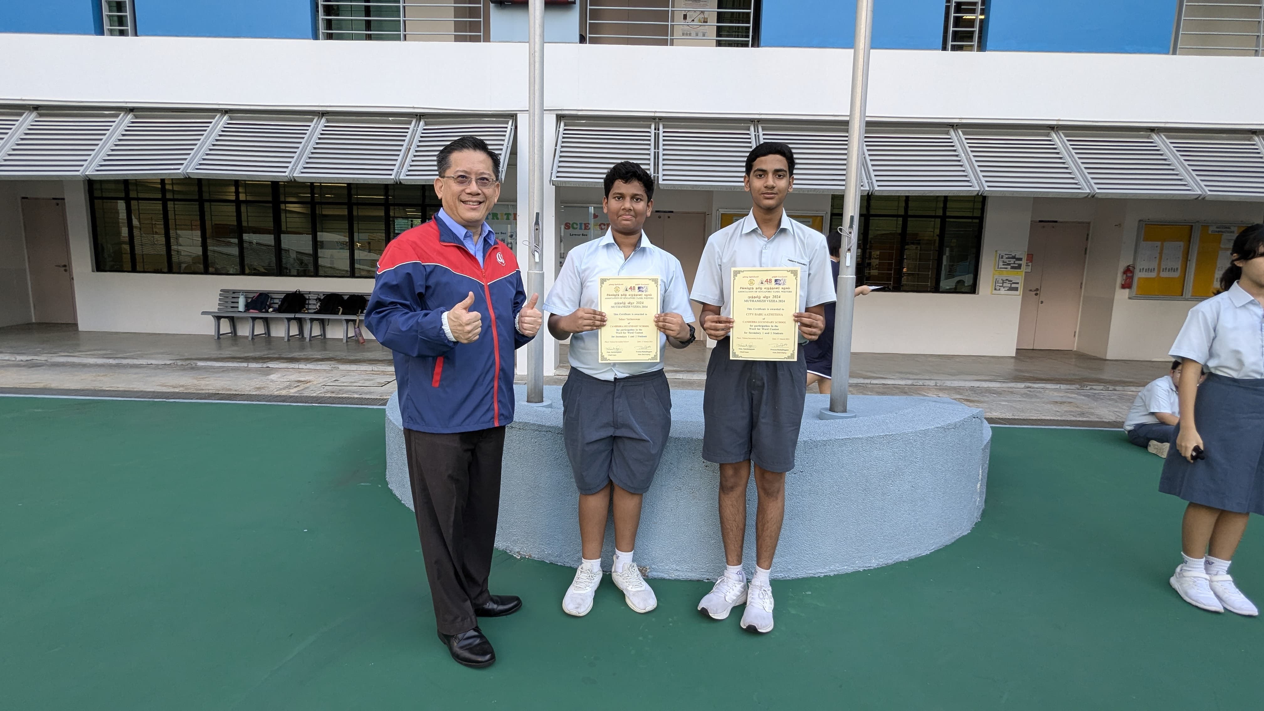 An adult giving thumbs up stands beside two students holding certificates in a school courtyard.