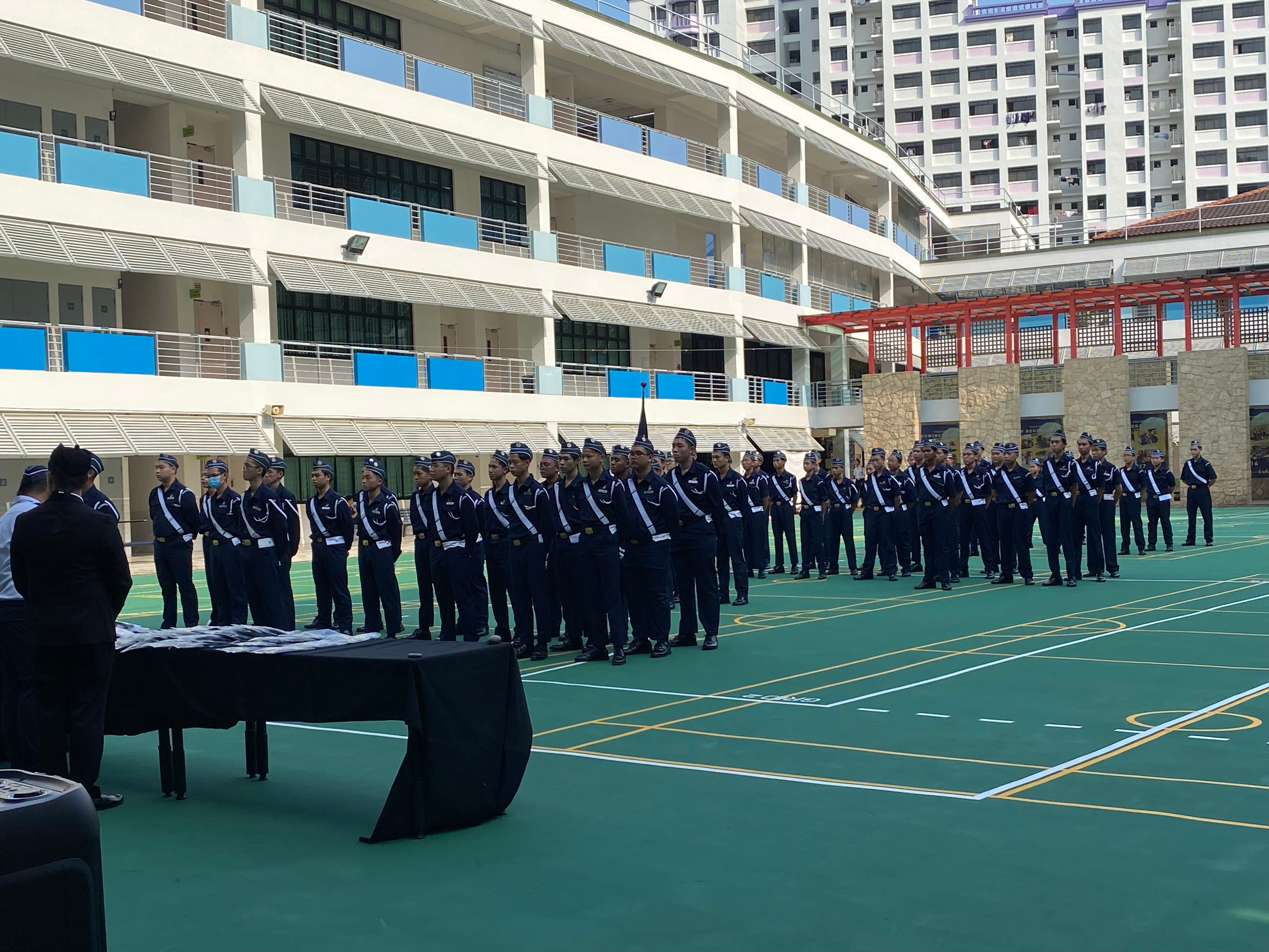 Rows of uniformed cadets in dark blue stand at attention on a green court in a school courtyard.