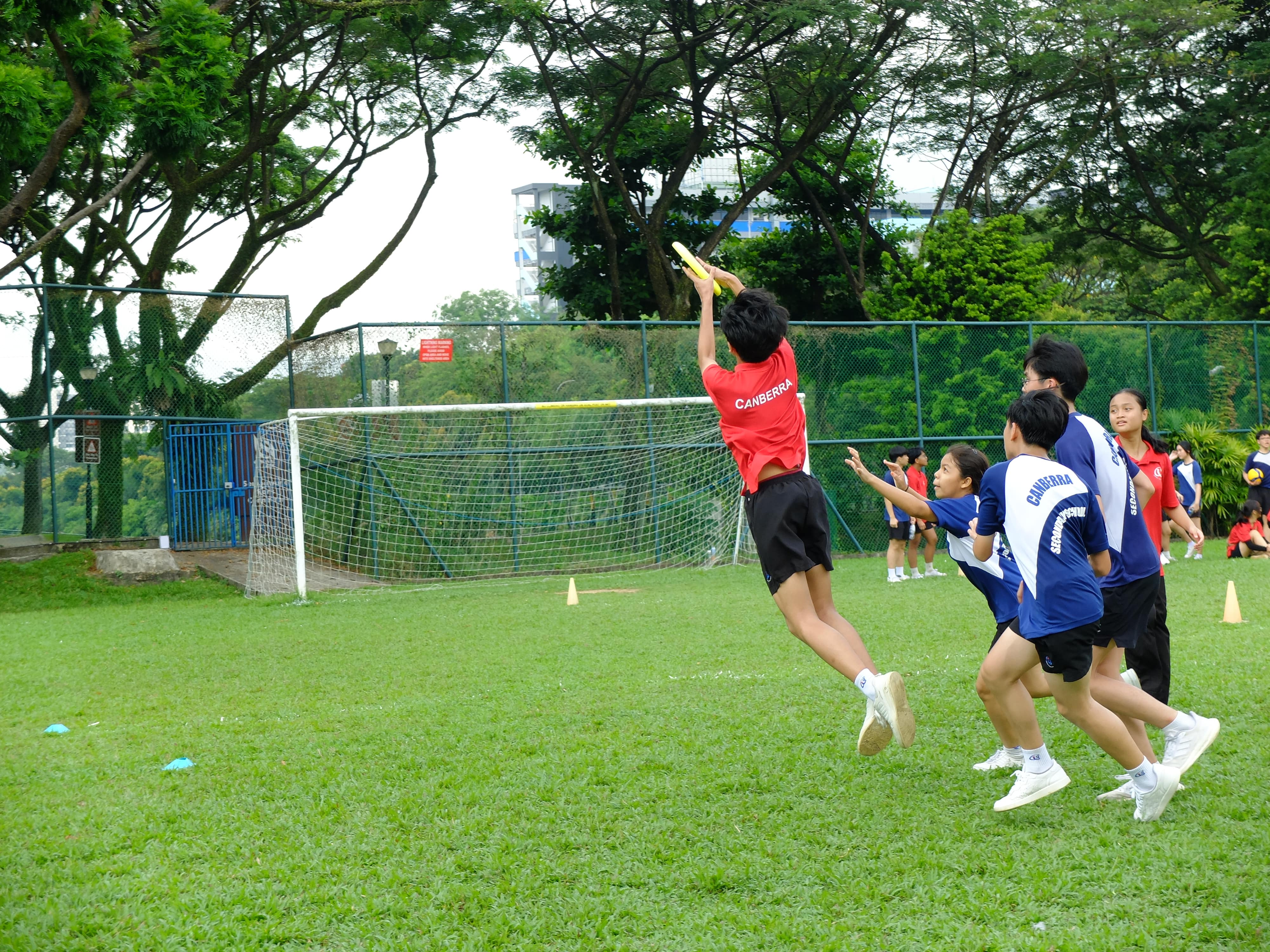 Students in red and blue Canberra jerseys play ultimate frisbee on a grass field near a soccer goal.