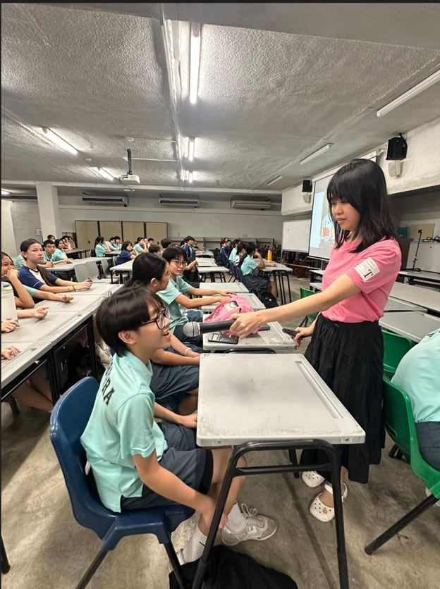 A teacher in a pink t-shirt and black skirt distributes materials to students seated at desks in a lecture-style classroom. Students are wearing light teal and dark blue school uniforms. A projector screen is visible in the background.