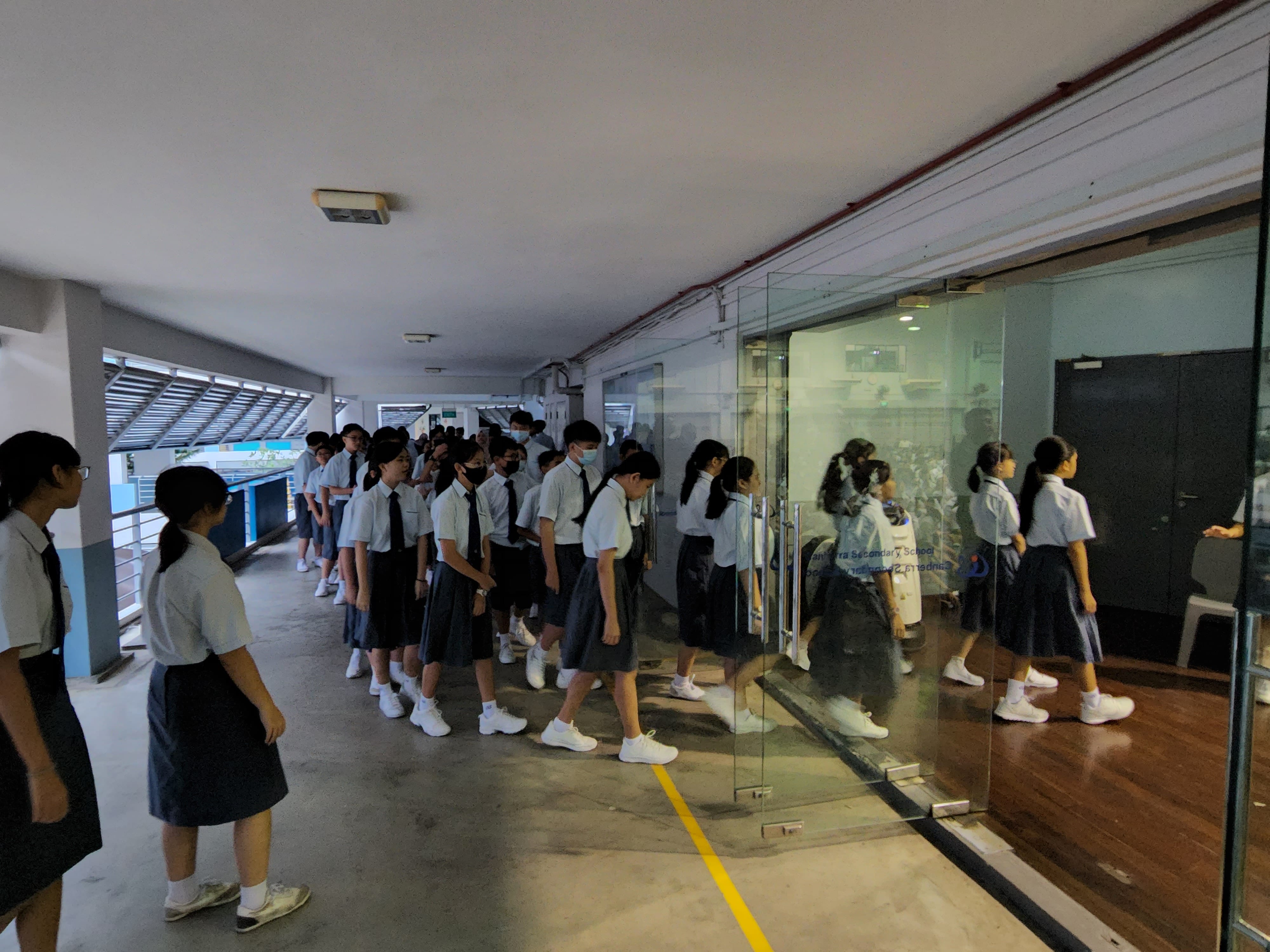 Students in white shirts and dark skirts queue in a school corridor beside a glass-walled room.