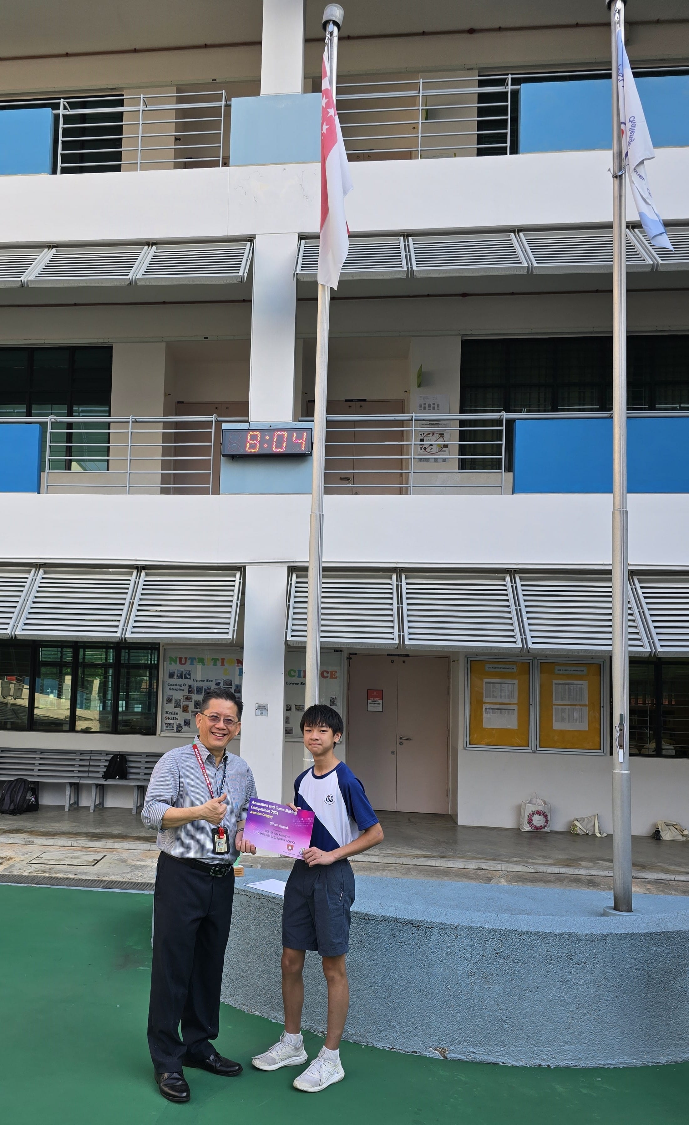 Two people stand near a flagpole outside a school building, holding a certificate together.