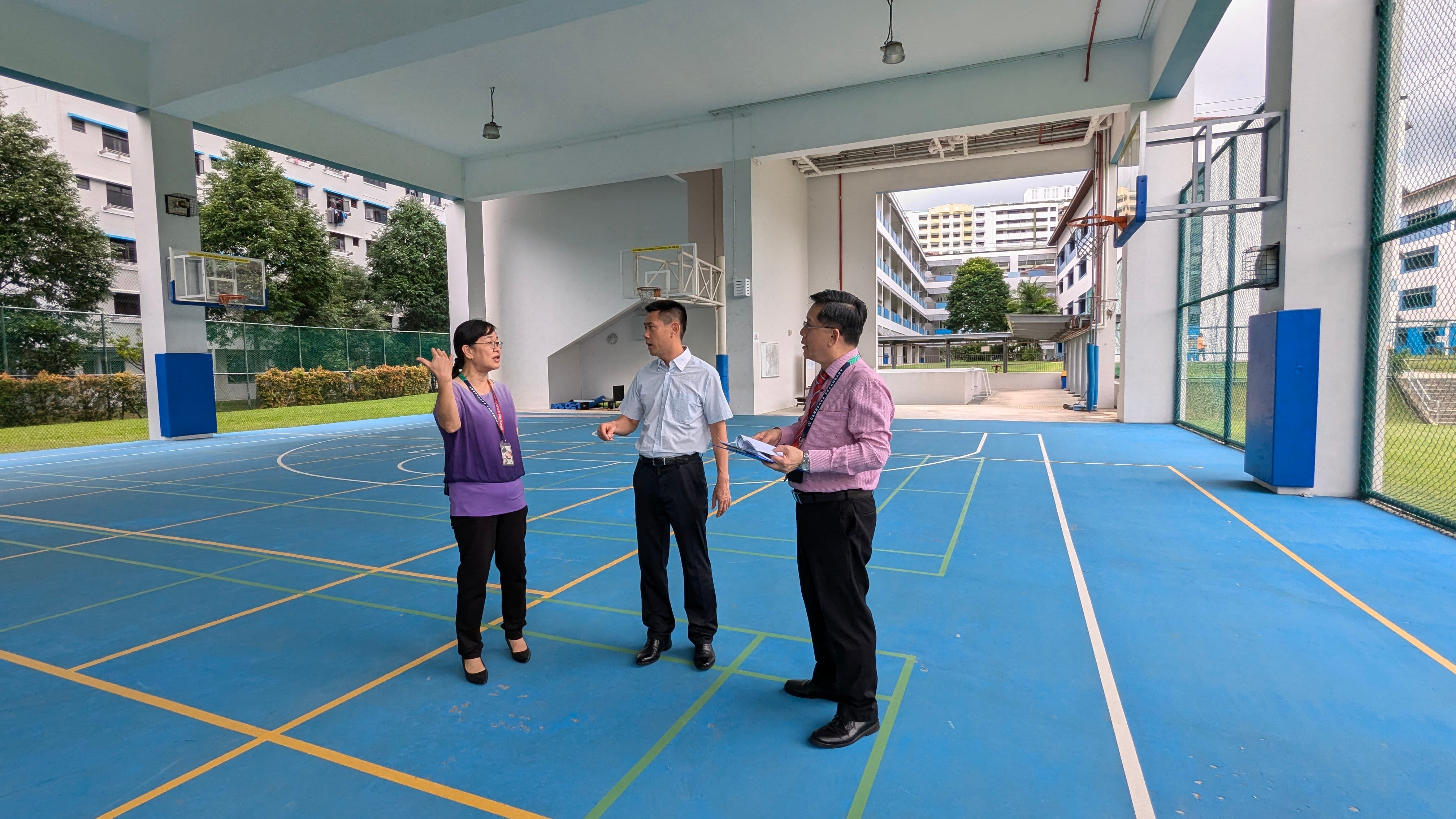Three people stand talking on a blue indoor basketball court with hoops and school buildings visible outside.