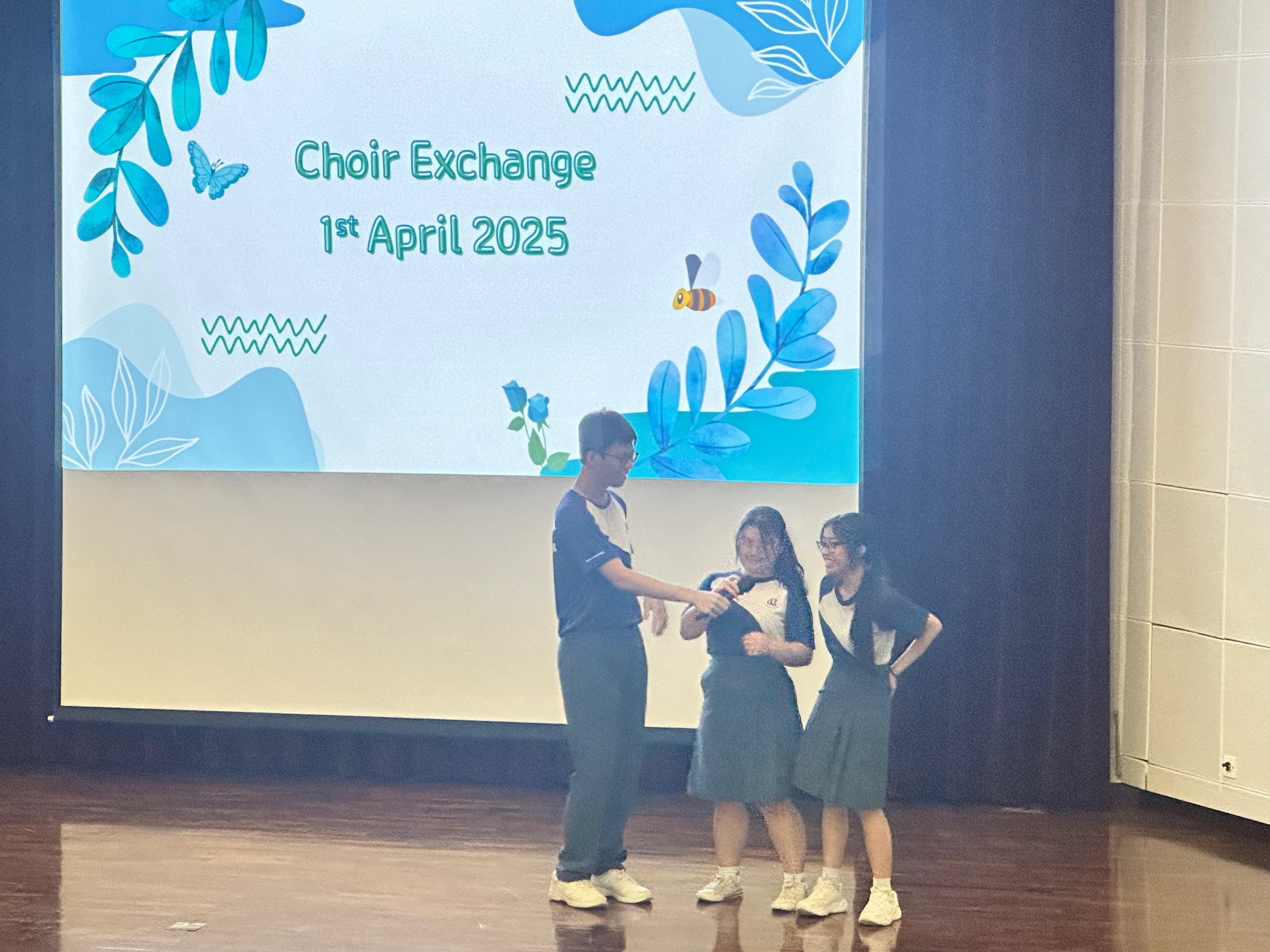 Three students stand on a stage in front of a projected slide reading Choir Exchange 1st April 2025.