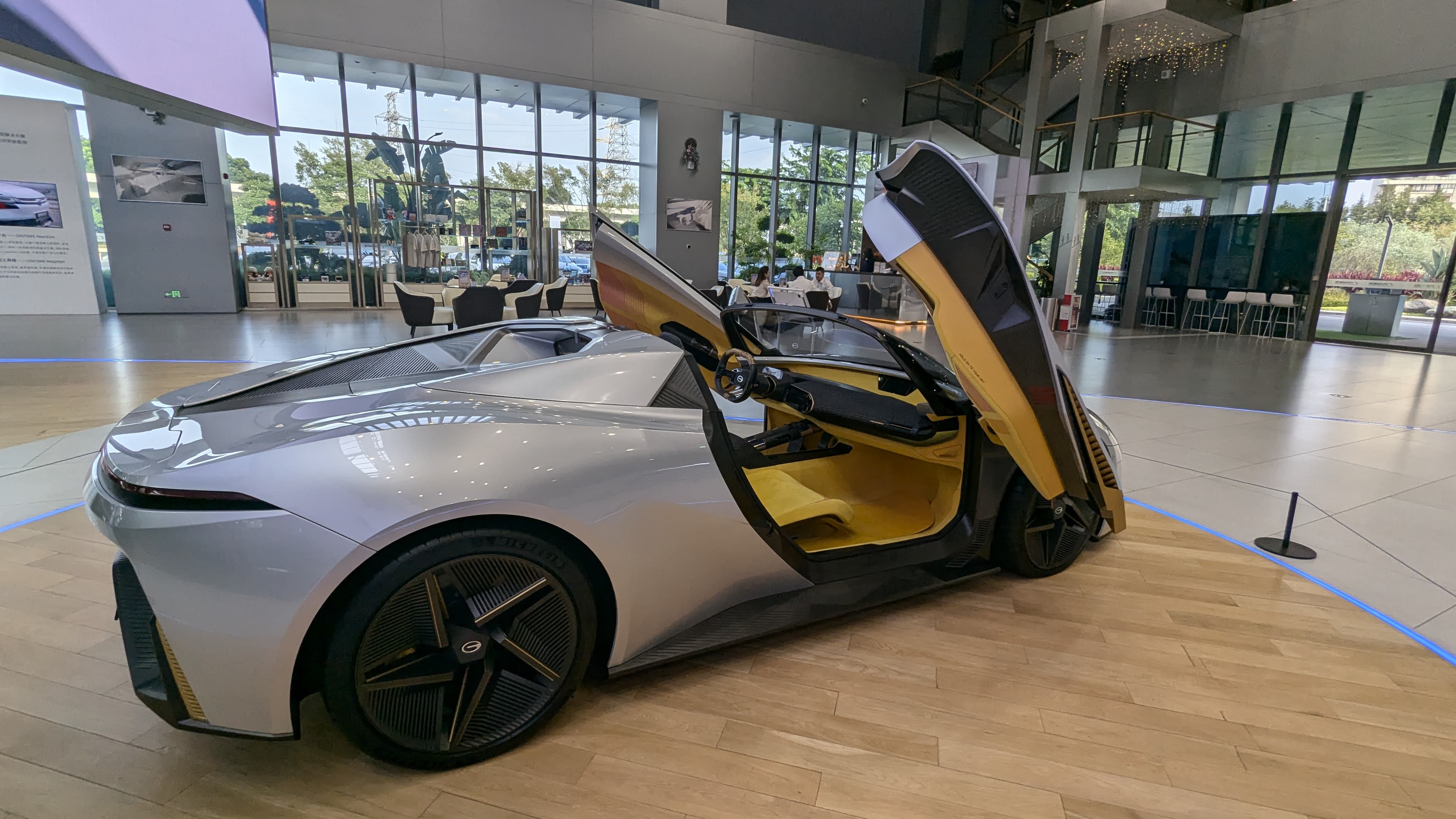 Silver concept sports car with butterfly doors open, revealing yellow interior, displayed in a modern showroom.