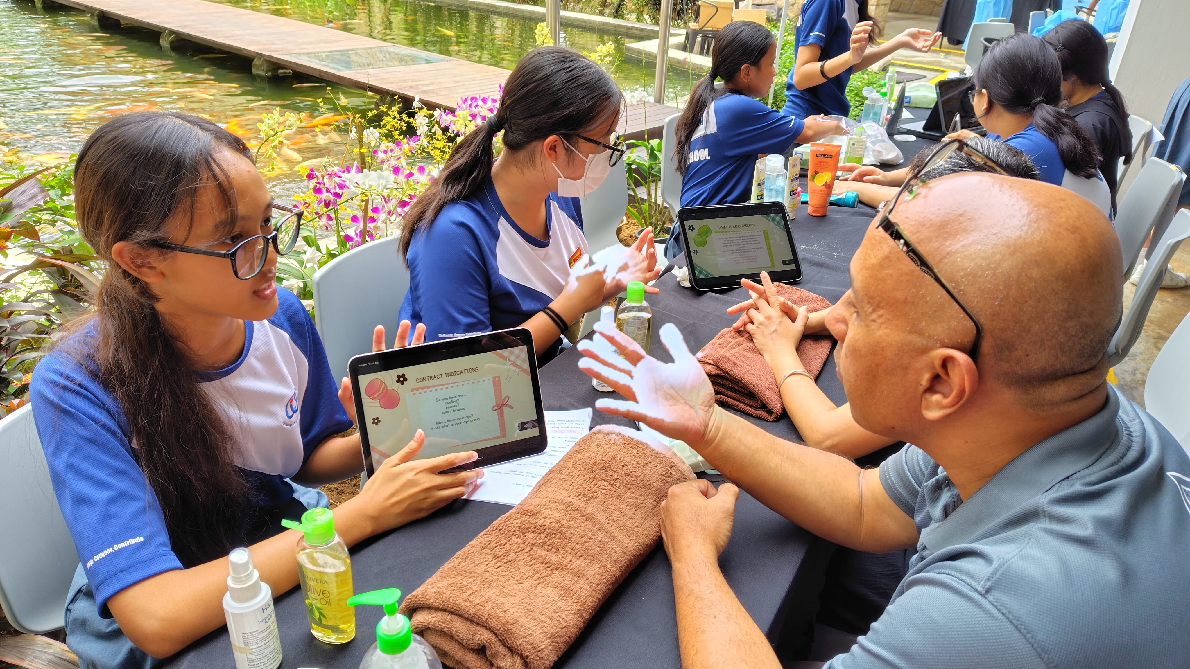 Two students in blue uniforms give a hand massage to a man at an outdoor table with tablets and lotions near a pond.