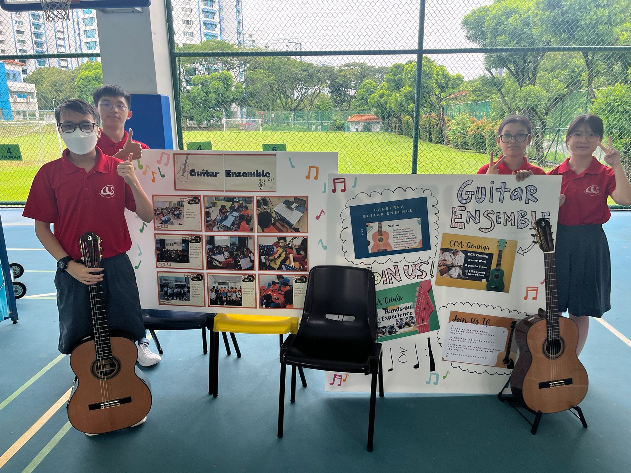 Four students in red uniforms stand beside Guitar Ensemble recruitment posters and two acoustic guitars on a school court.