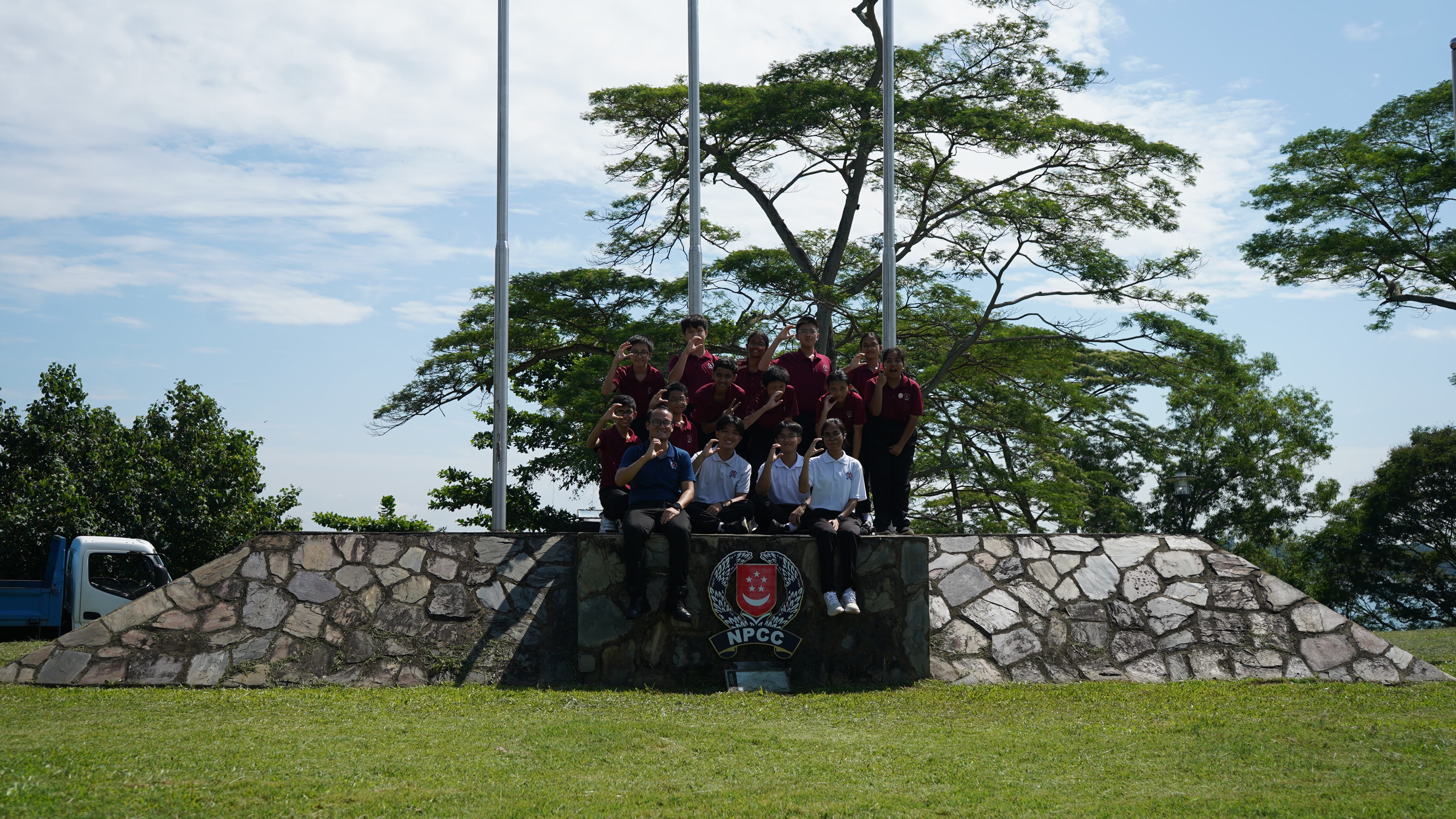 Group of students posing on a stone platform with flagpoles, trees and blue sky behind.