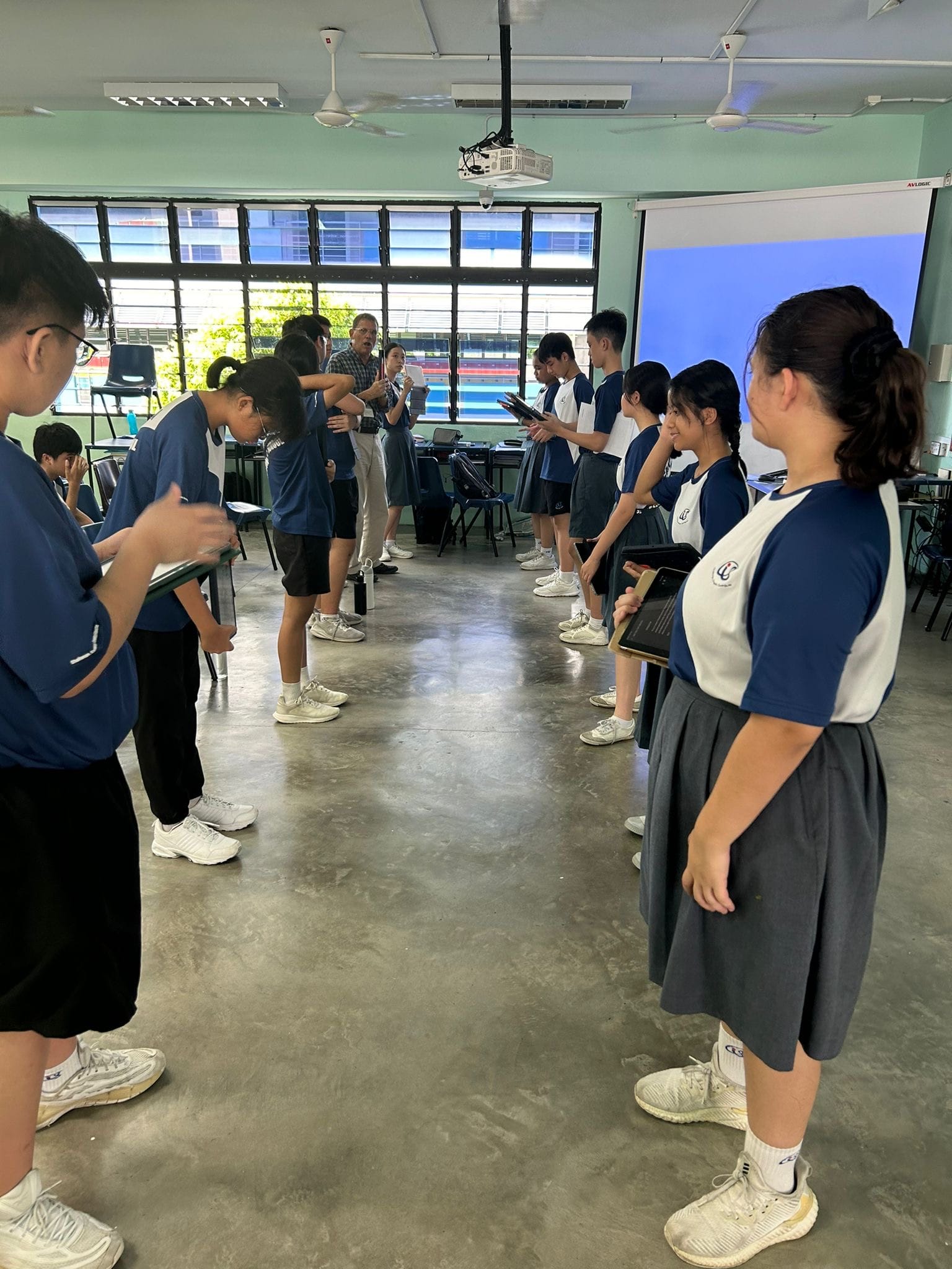 Students in blue uniforms stand in two facing rows in a classroom while a teacher speaks at the front.