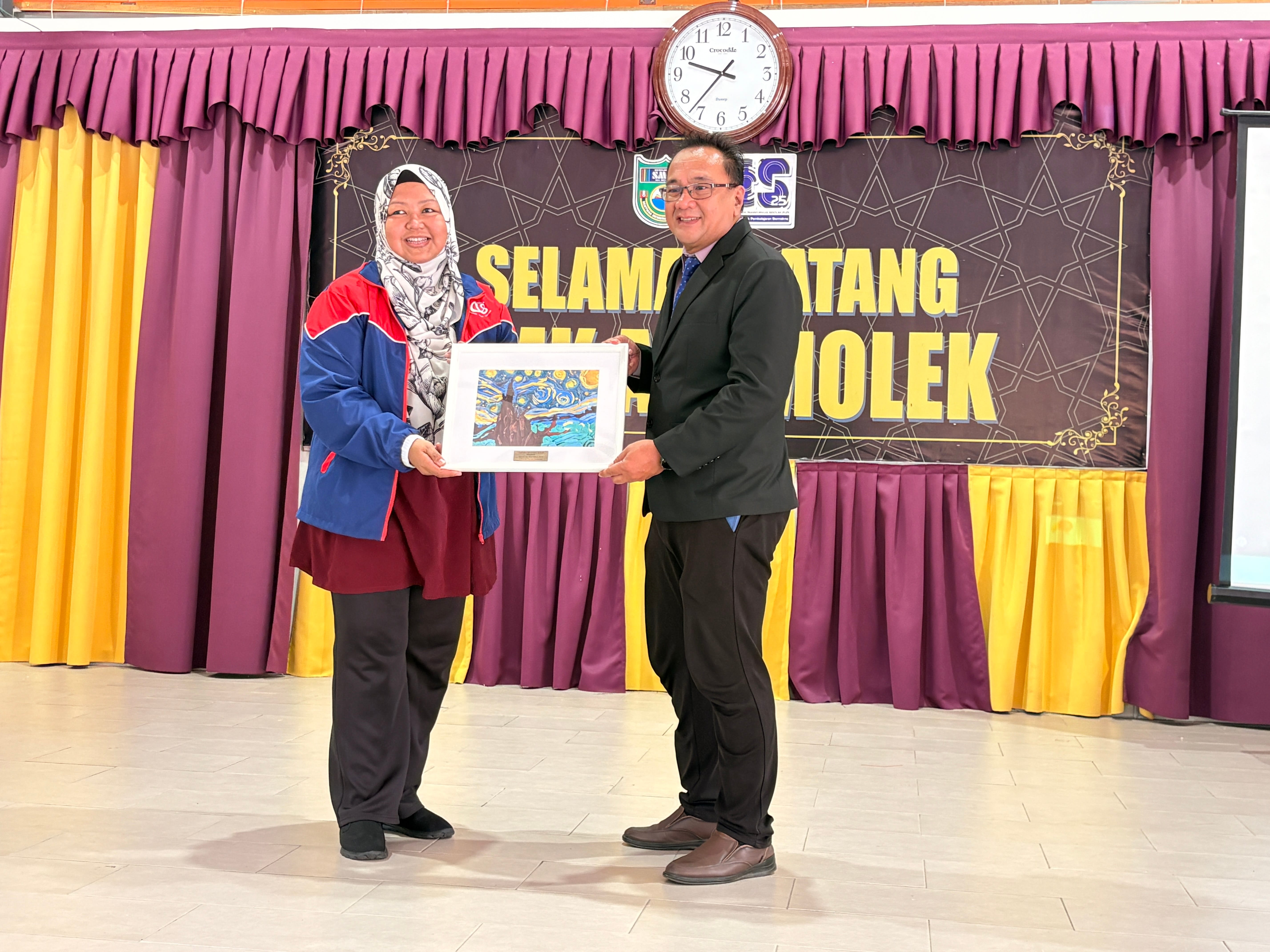 Two people hold a framed painting together in front of a banner reading "Selamat Datang" in a decorated hall.