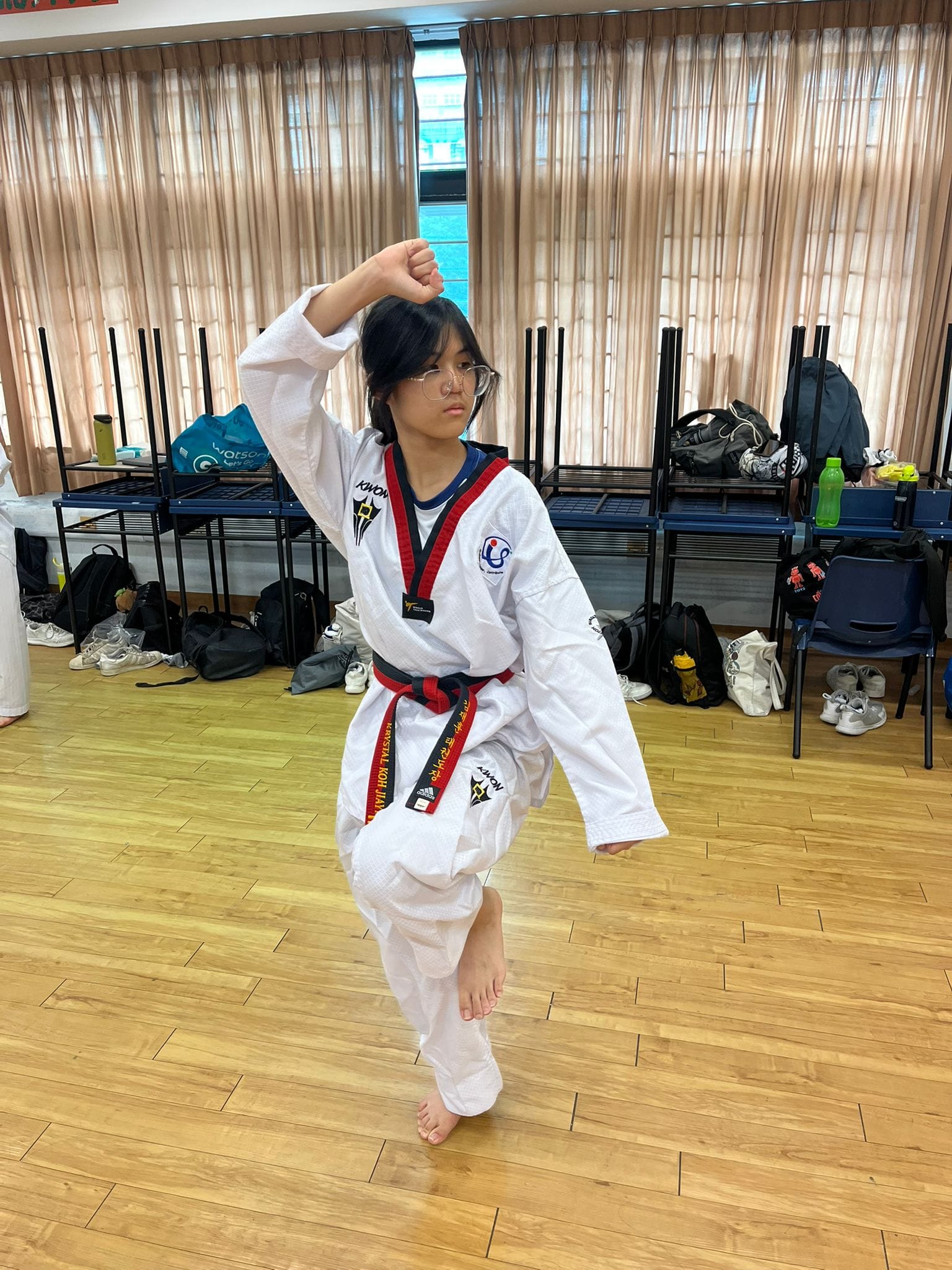 A young girl in a white taekwondo uniform with a red and black belt performs a pose on a wooden floor.