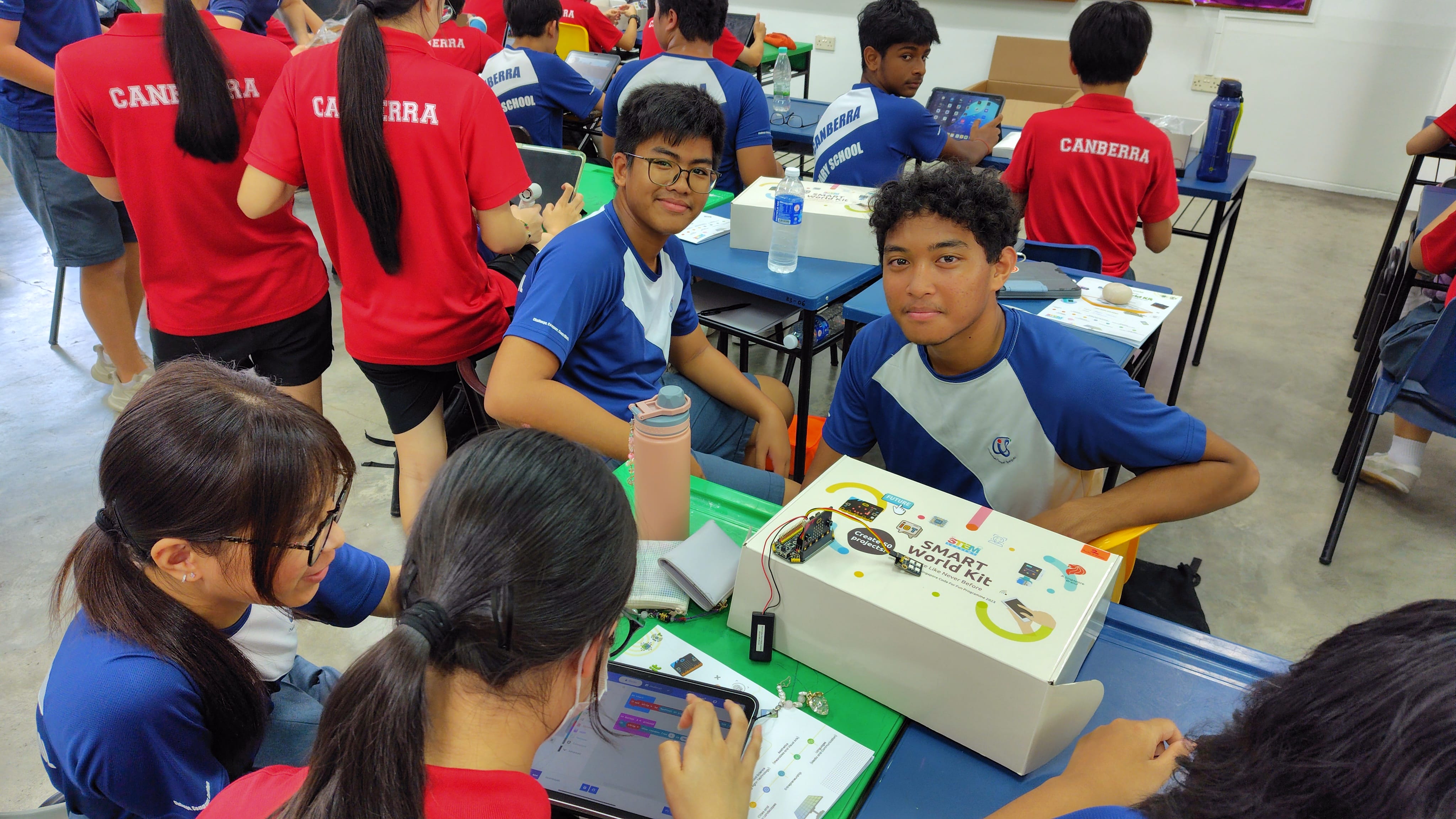 Students in blue and red uniforms work with a STEM Smart World Kit and tablets in a classroom.
