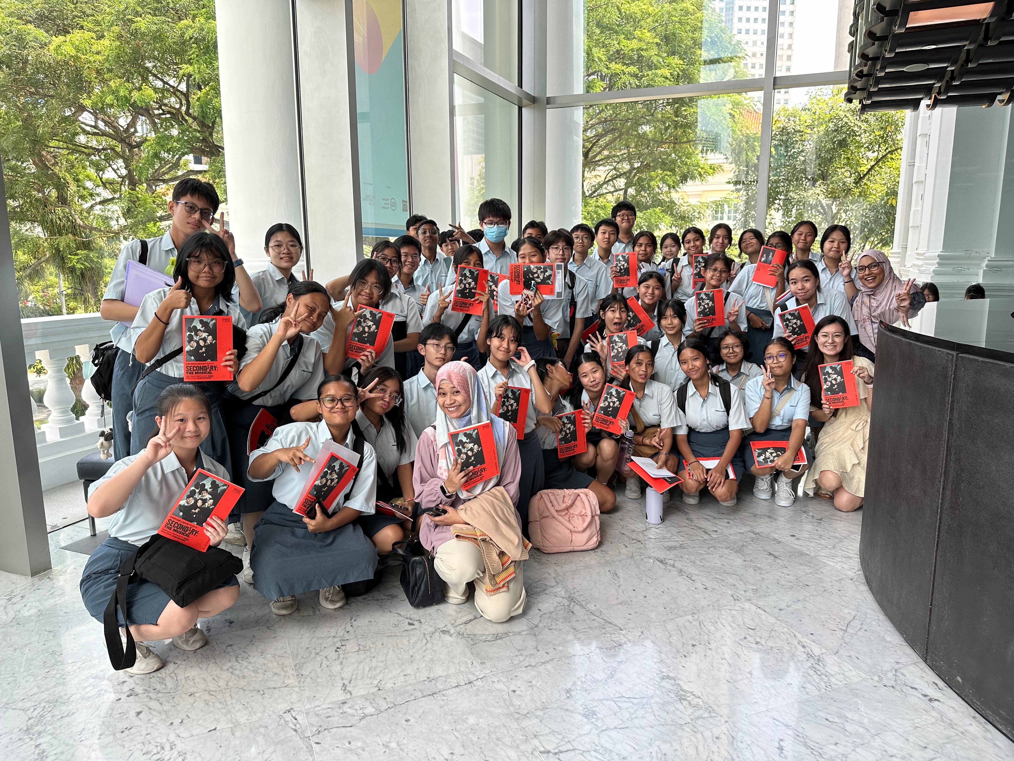 A large group of uniformed students poses indoors holding red playbills, smiling and making peace signs.