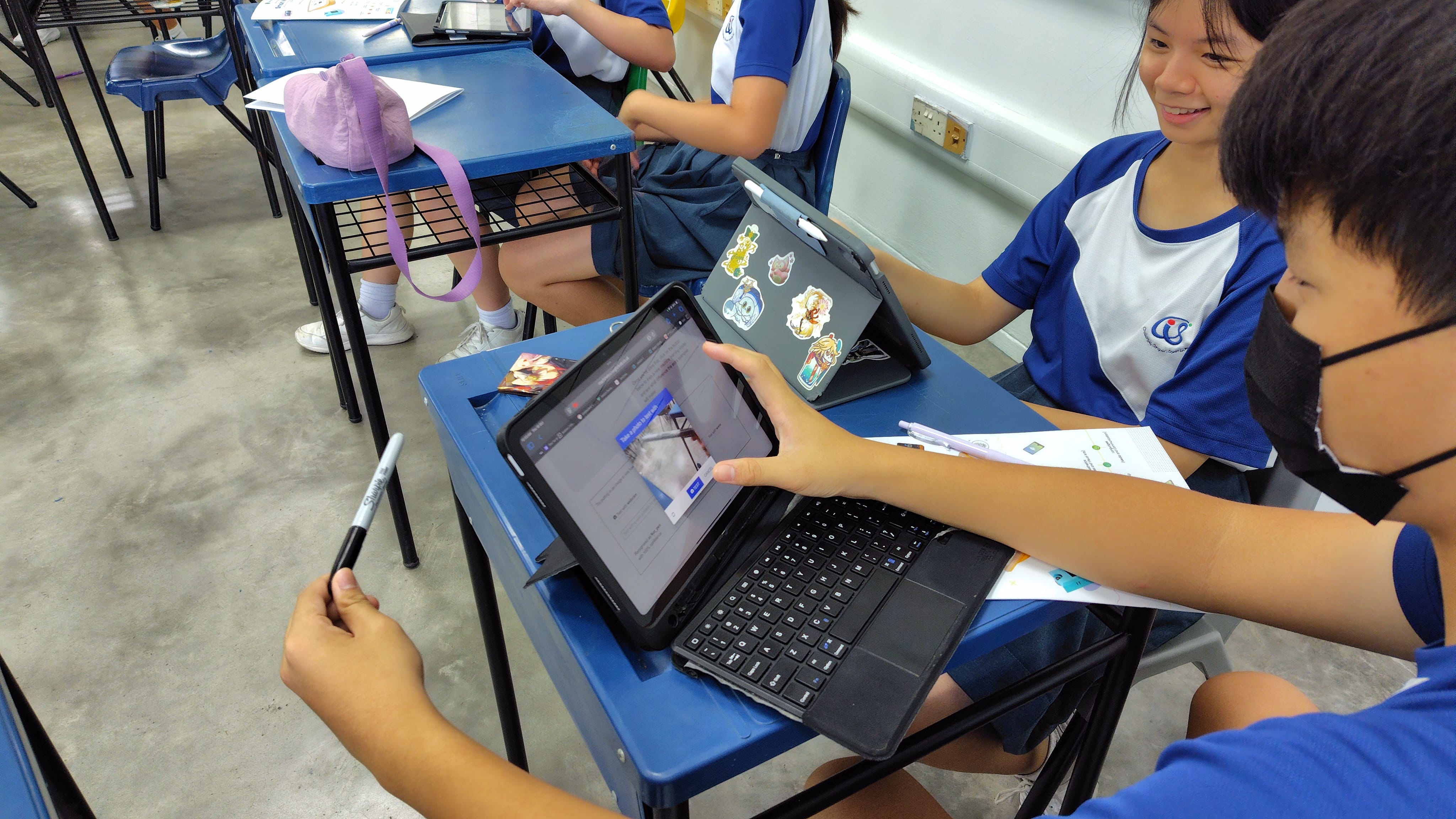 Two students in blue uniforms sit at school desks, sharing a tablet with a keyboard case in a classroom.