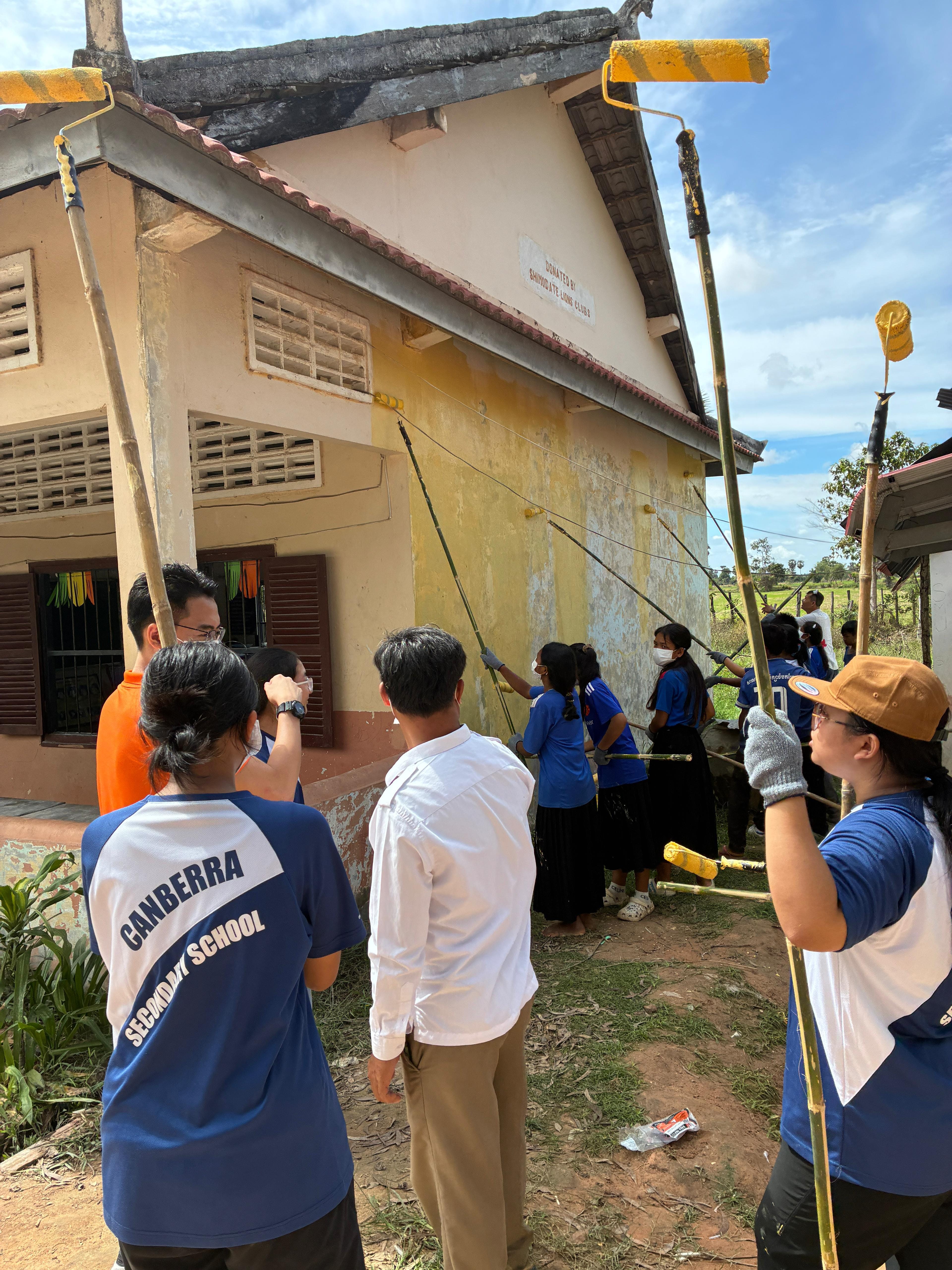 Canberra Secondary School volunteers painting yellow on the exterior wall of a rural building using long-handled rollers.