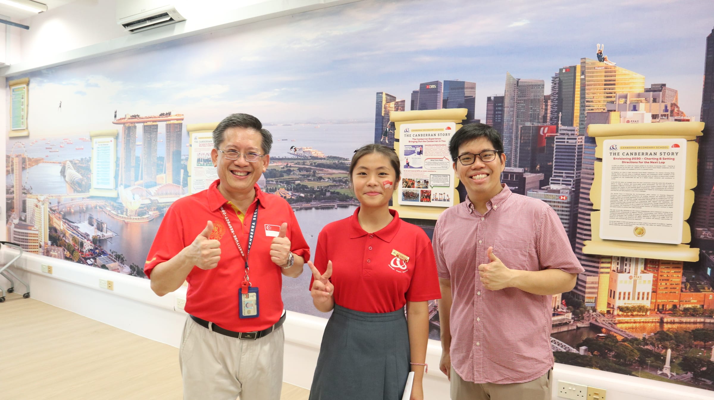 Three smiling people in red shirts give thumbs up before a large Singapore cityscape mural at Canberra Secondary School.