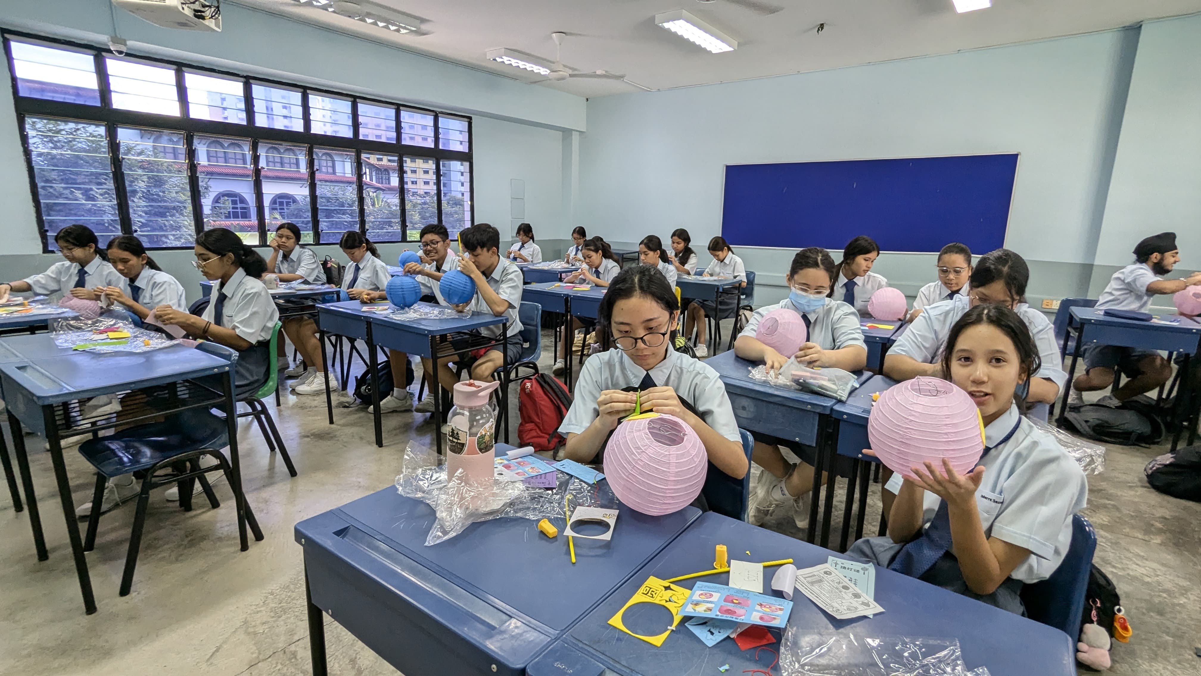 Students in school uniforms sit at blue desks assembling colorful paper lanterns in a classroom.