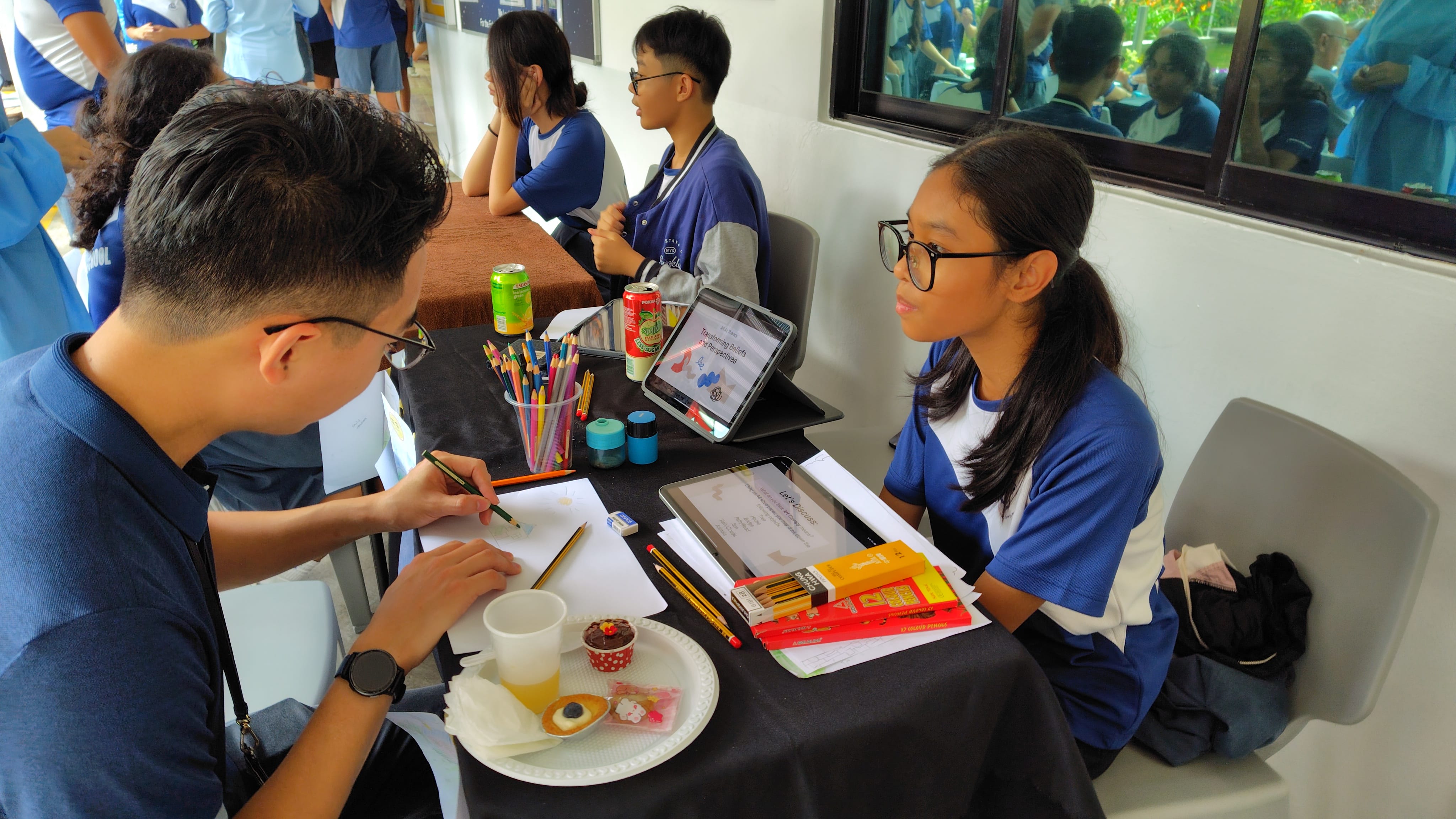 Two students in blue uniforms sit at a table with pencils, a tablet, snacks, and papers during a school activity.