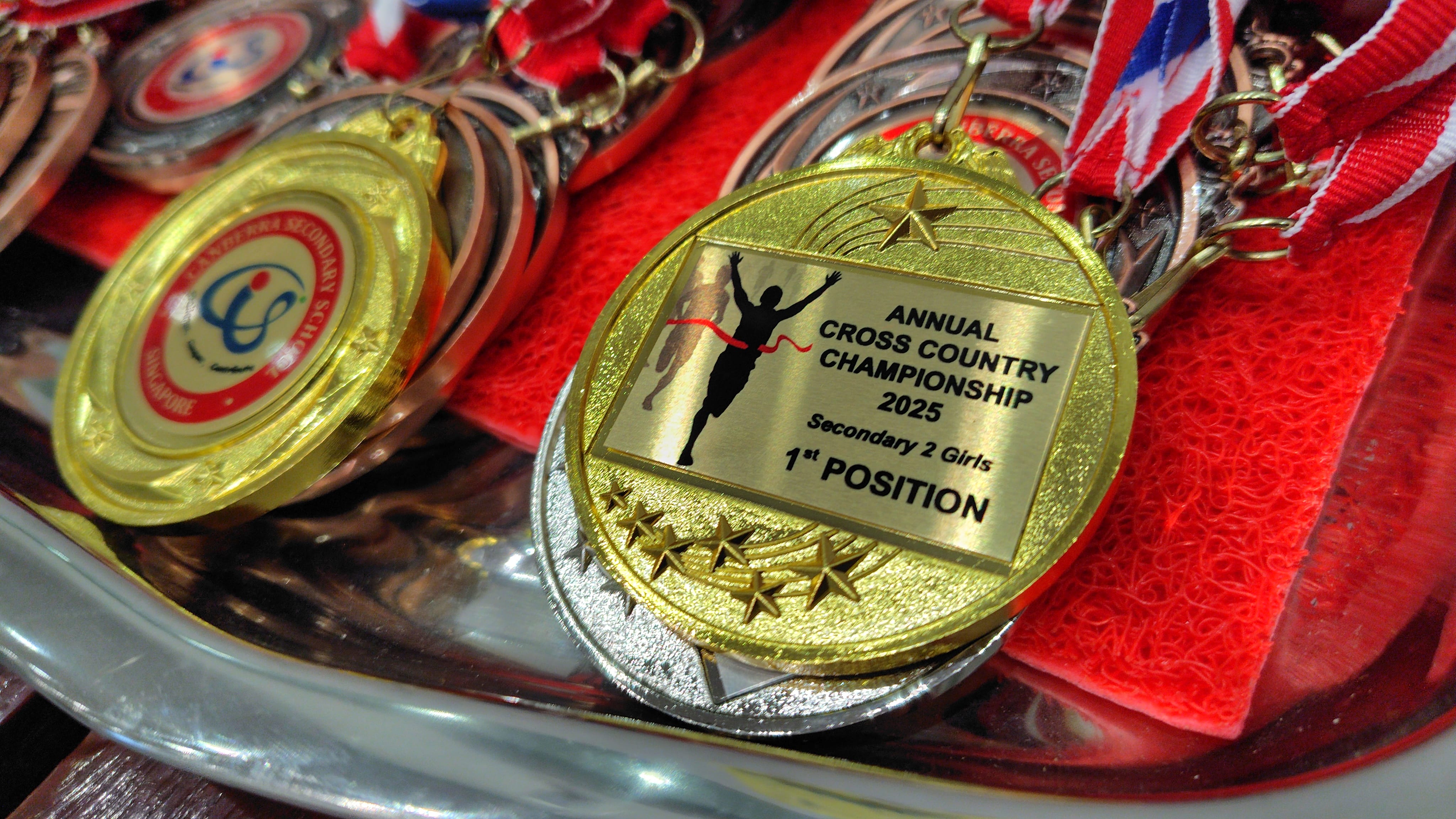 Gold medal reading Annual Cross Country Championship 2025 Secondary 2 Girls 1st Position, with red ribbons nearby.