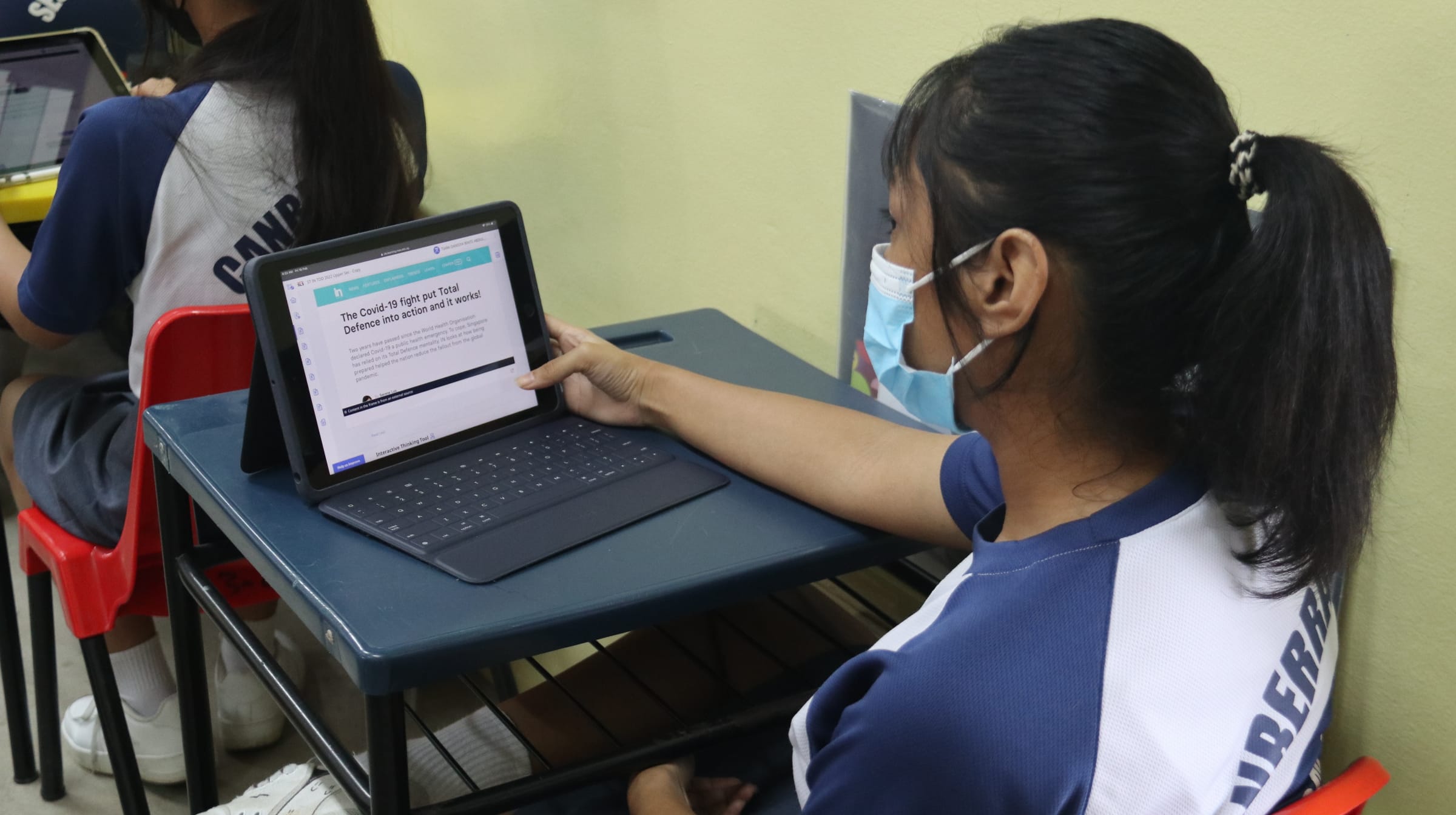 A masked student in school uniform touches a tablet screen showing a Covid-19 and Total Defence article at a desk.