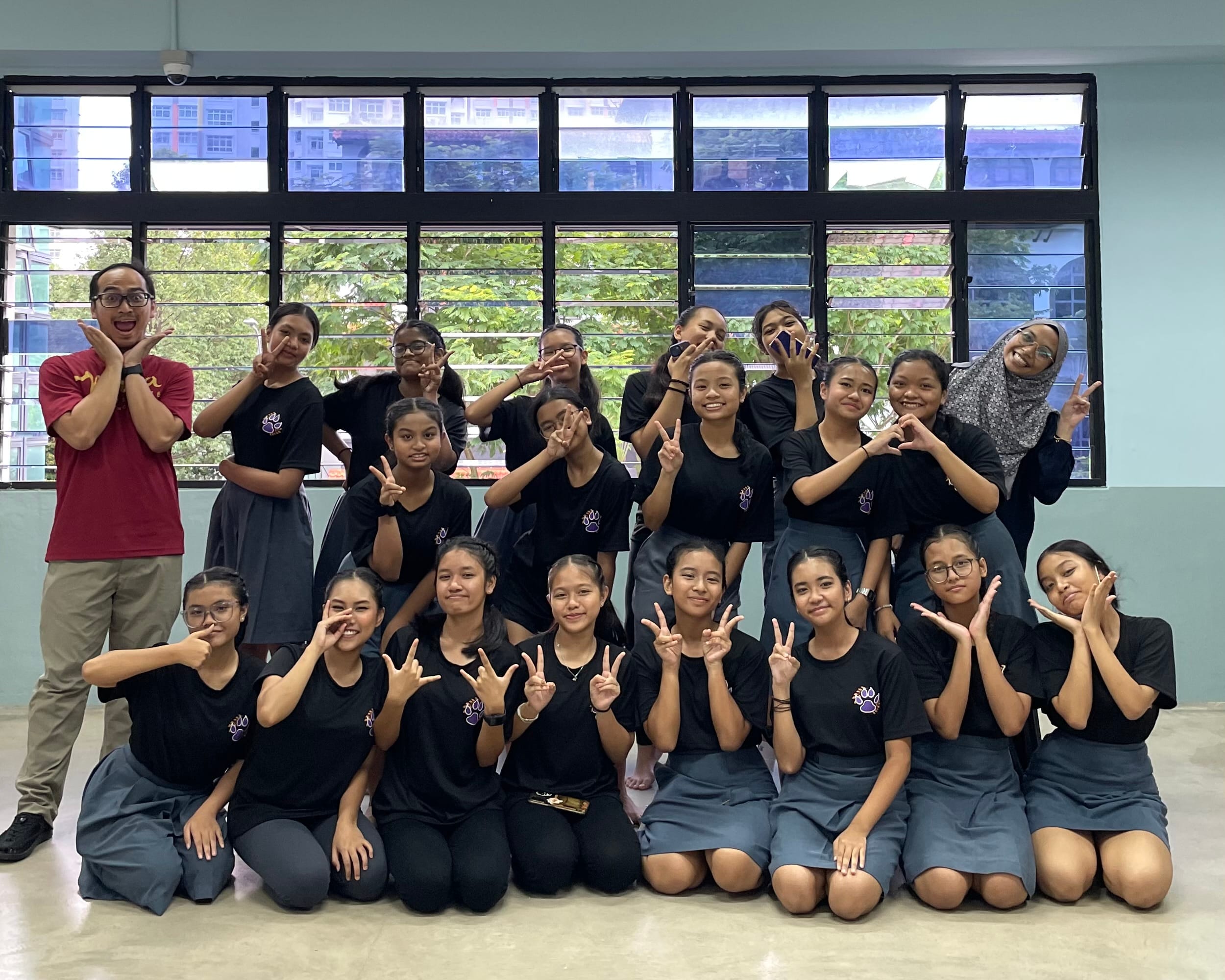 A group of teenage girls in black shirts pose with peace signs alongside a male teacher in a school hall.