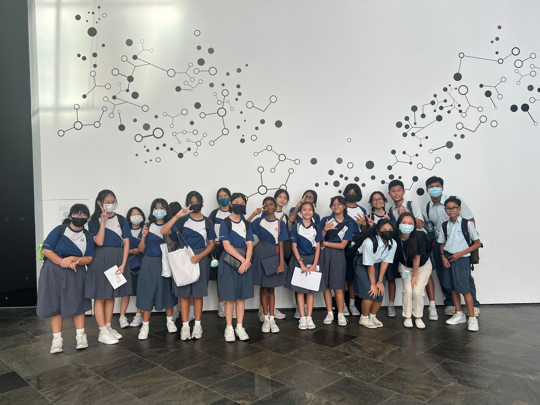 A group of students in blue and grey school uniforms pose in front of a white wall with molecular dot patterns.