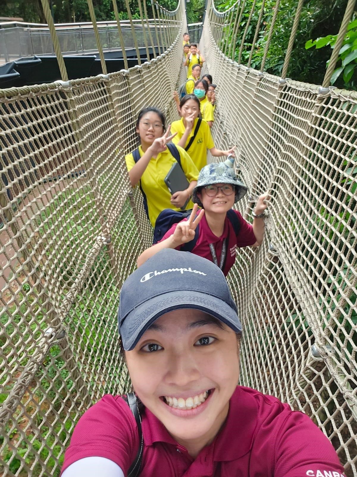 A woman in a Champion cap takes a selfie on a rope suspension bridge with a group of students in yellow shirts behind her.