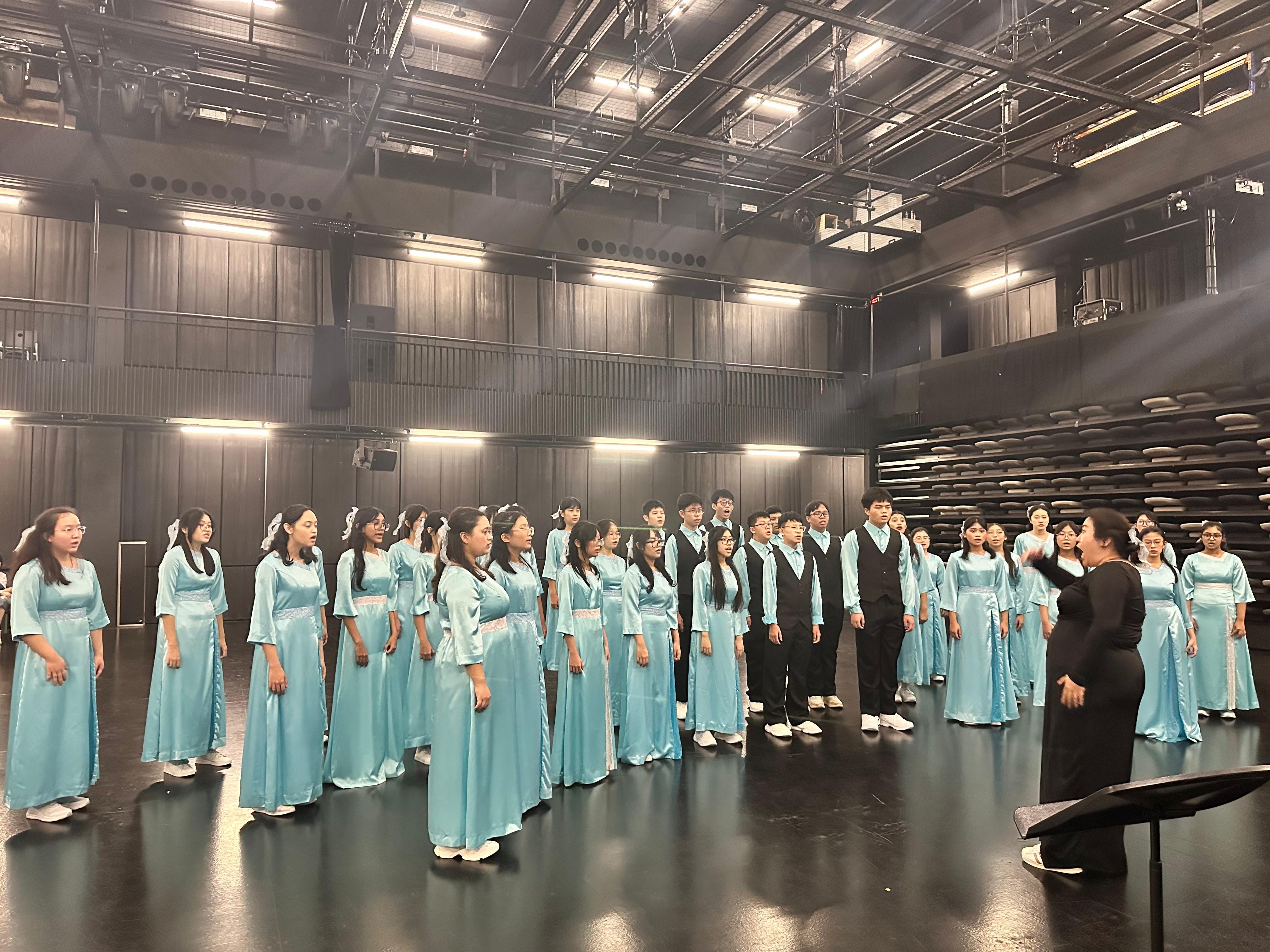 A choir in light blue satin gowns and black vests performs on a dark stage, led by a conductor in black.