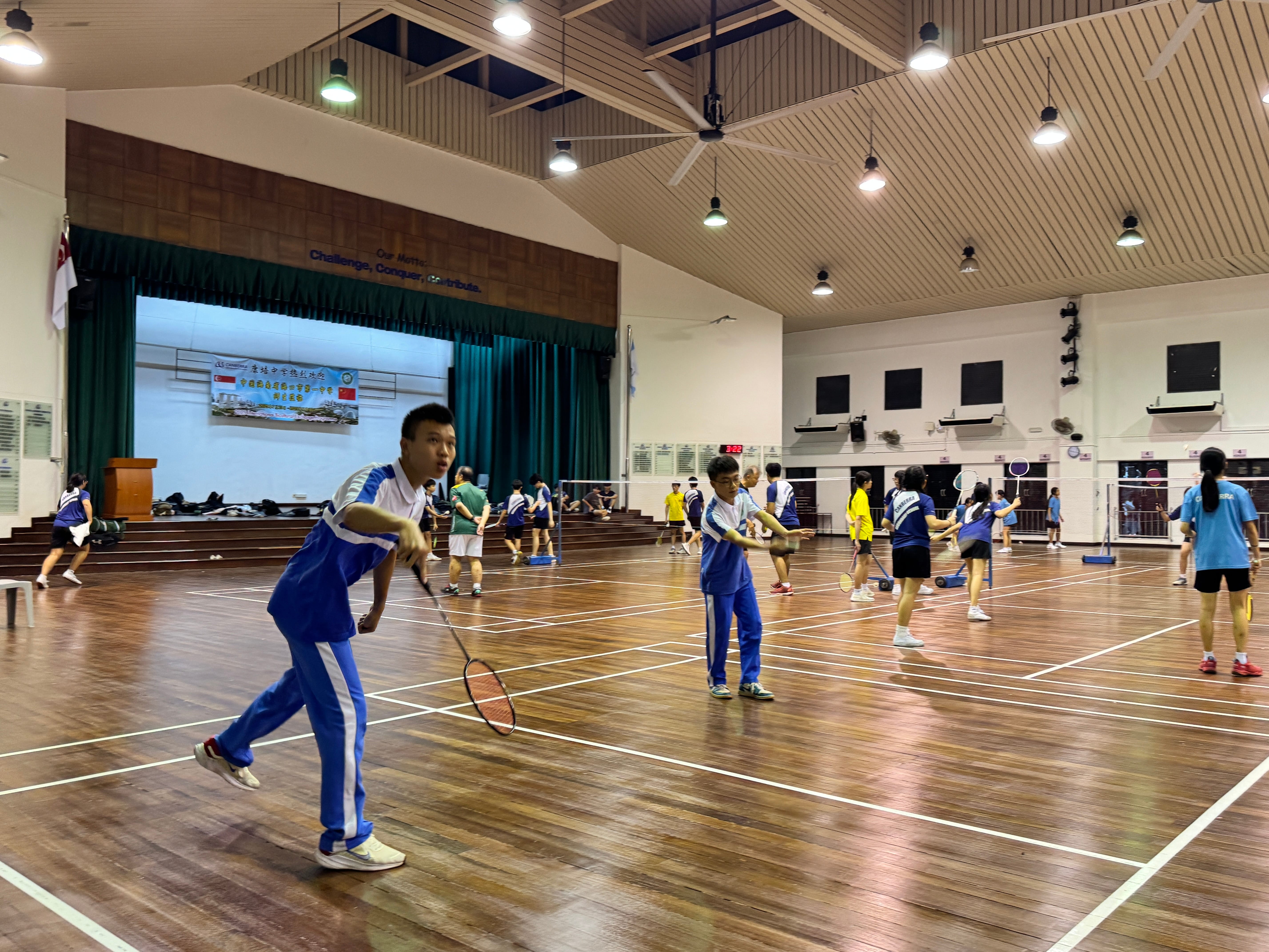 Students play badminton on multiple courts in a large indoor gym, with a boy serving in the foreground.