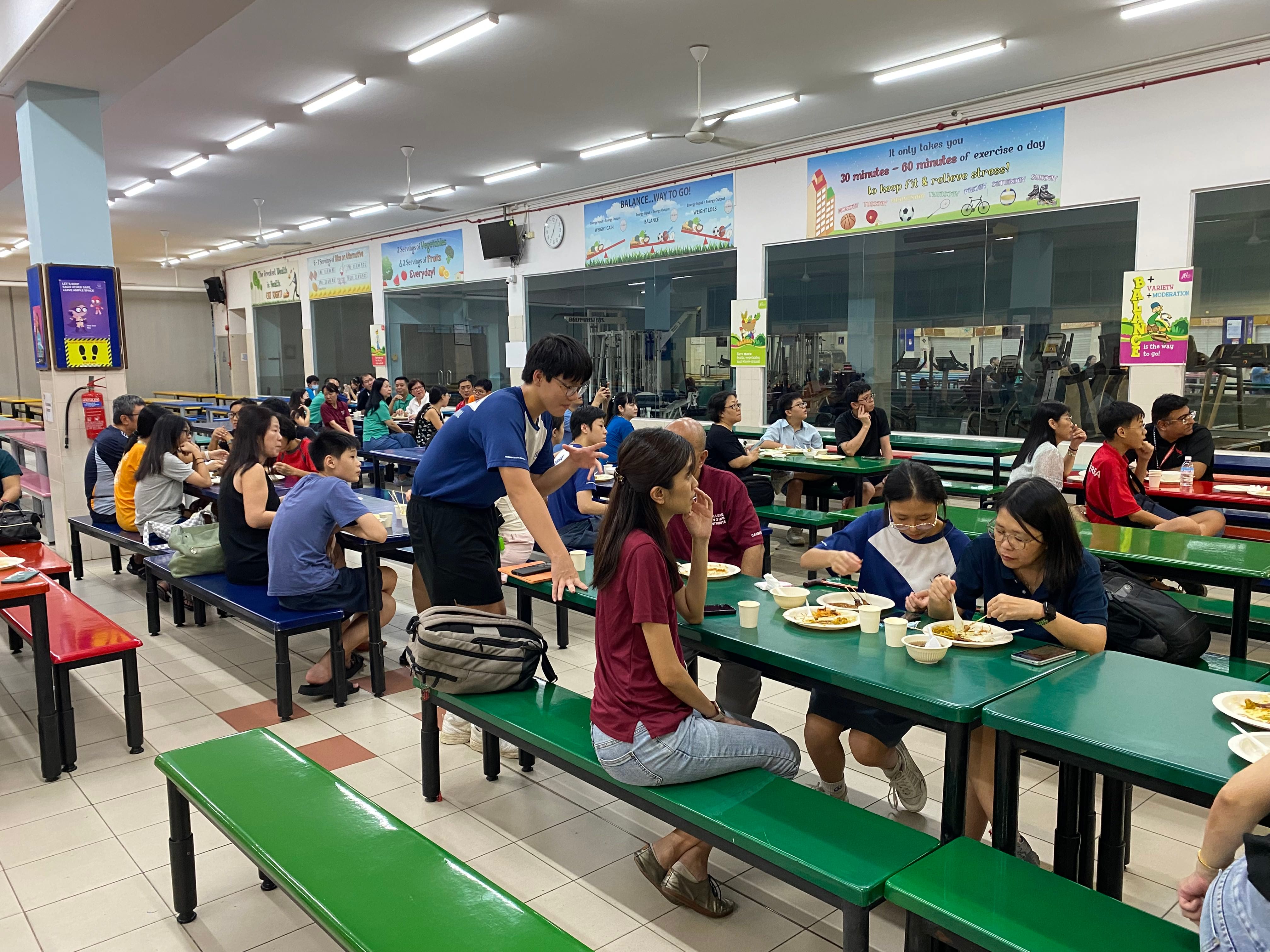 A busy school canteen with many people sitting at green benches eating meals, health posters on the walls.