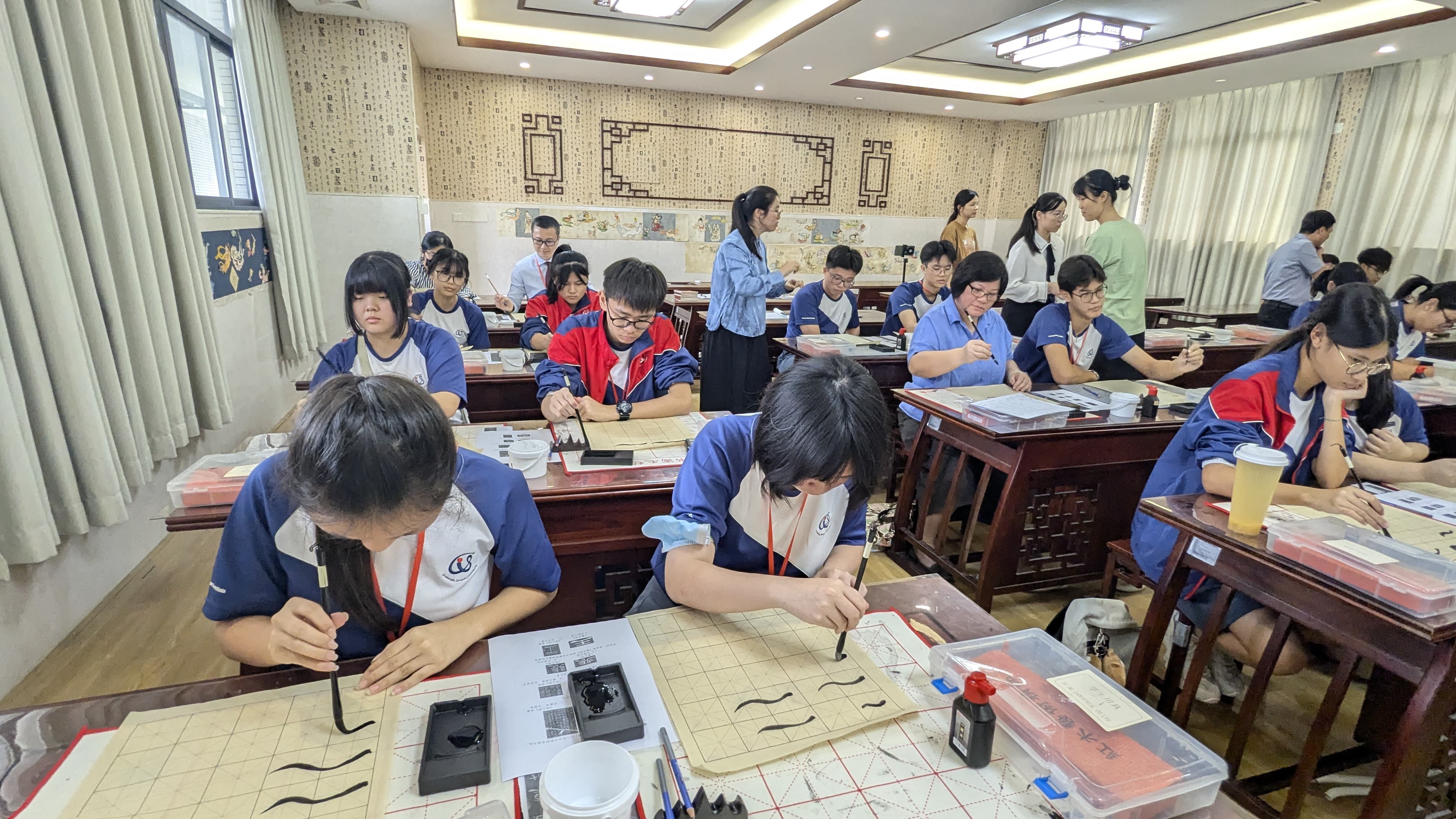 Students in blue uniforms practice Chinese calligraphy with brushes and ink at wooden desks in a decorated classroom.