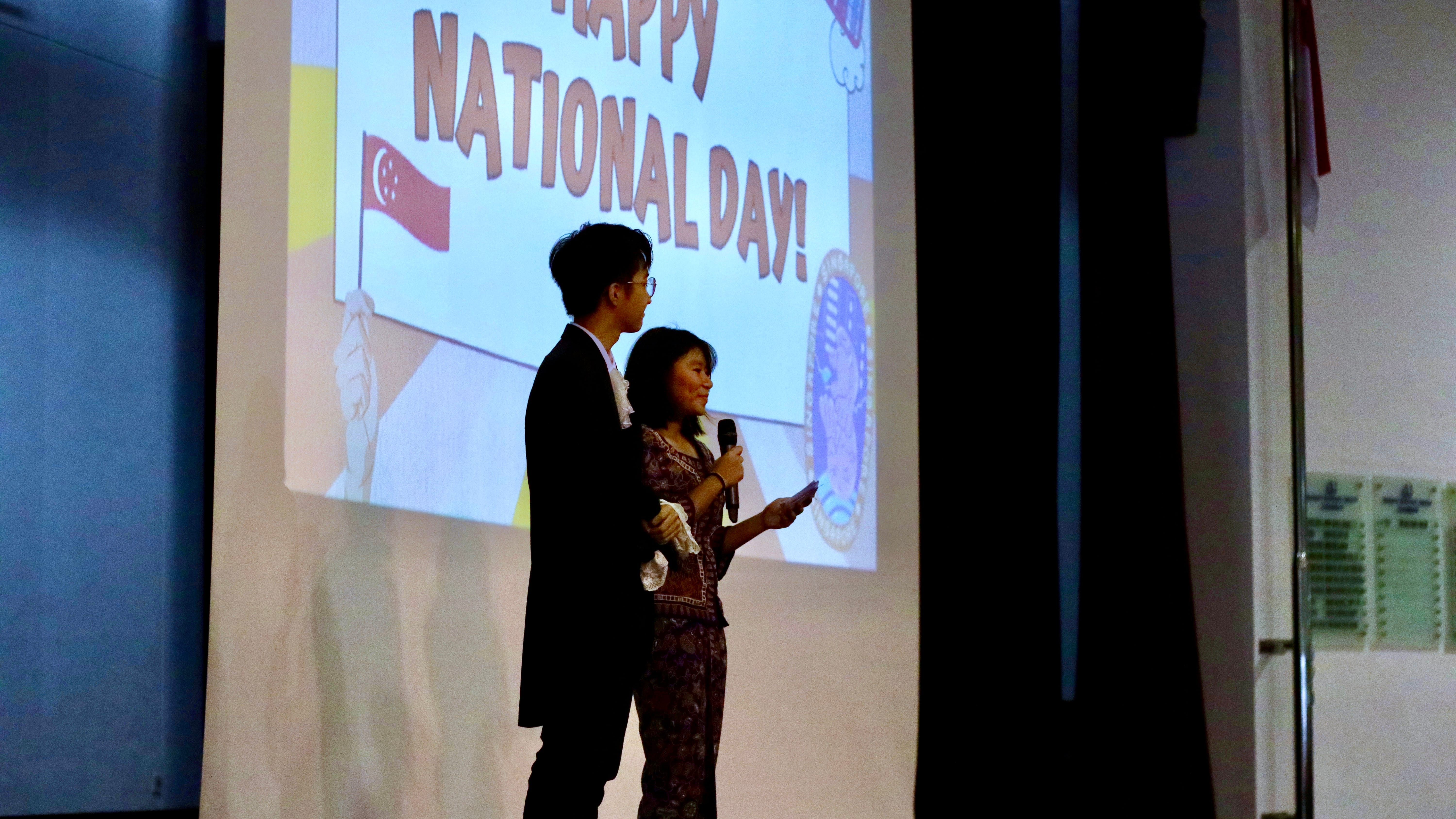 Two presenters on stage; woman speaks into microphone before a screen reading Happy National Day! with a flag.
