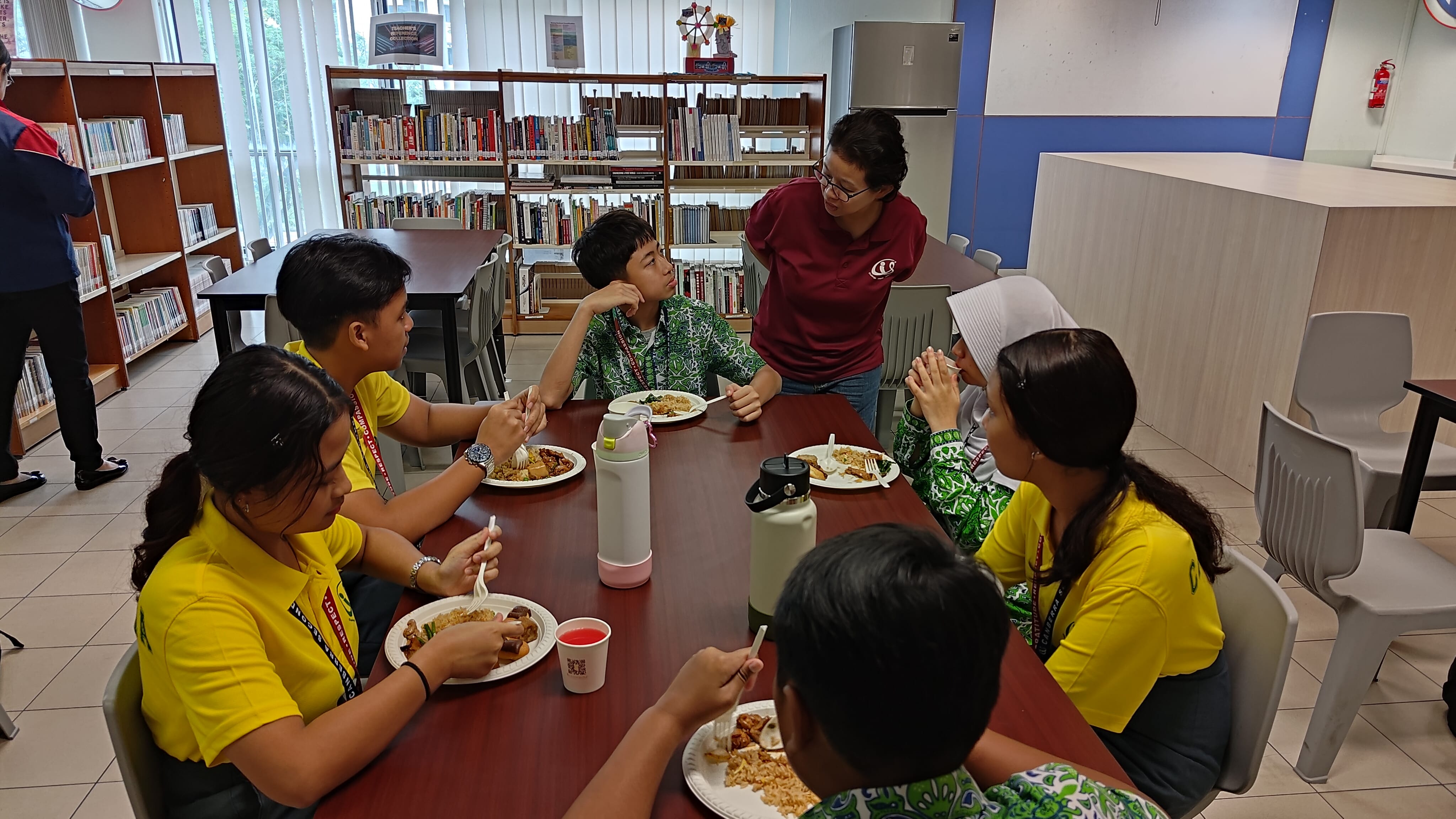 Several students in yellow shirts eating meals at a table in a library while an adult in maroon speaks to them.