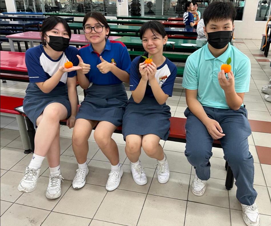 Four secondary school students sitting on a bench in the school canteen, each holding a mandarin orange and smiling at the camera during a Lunar New Year celebration.
