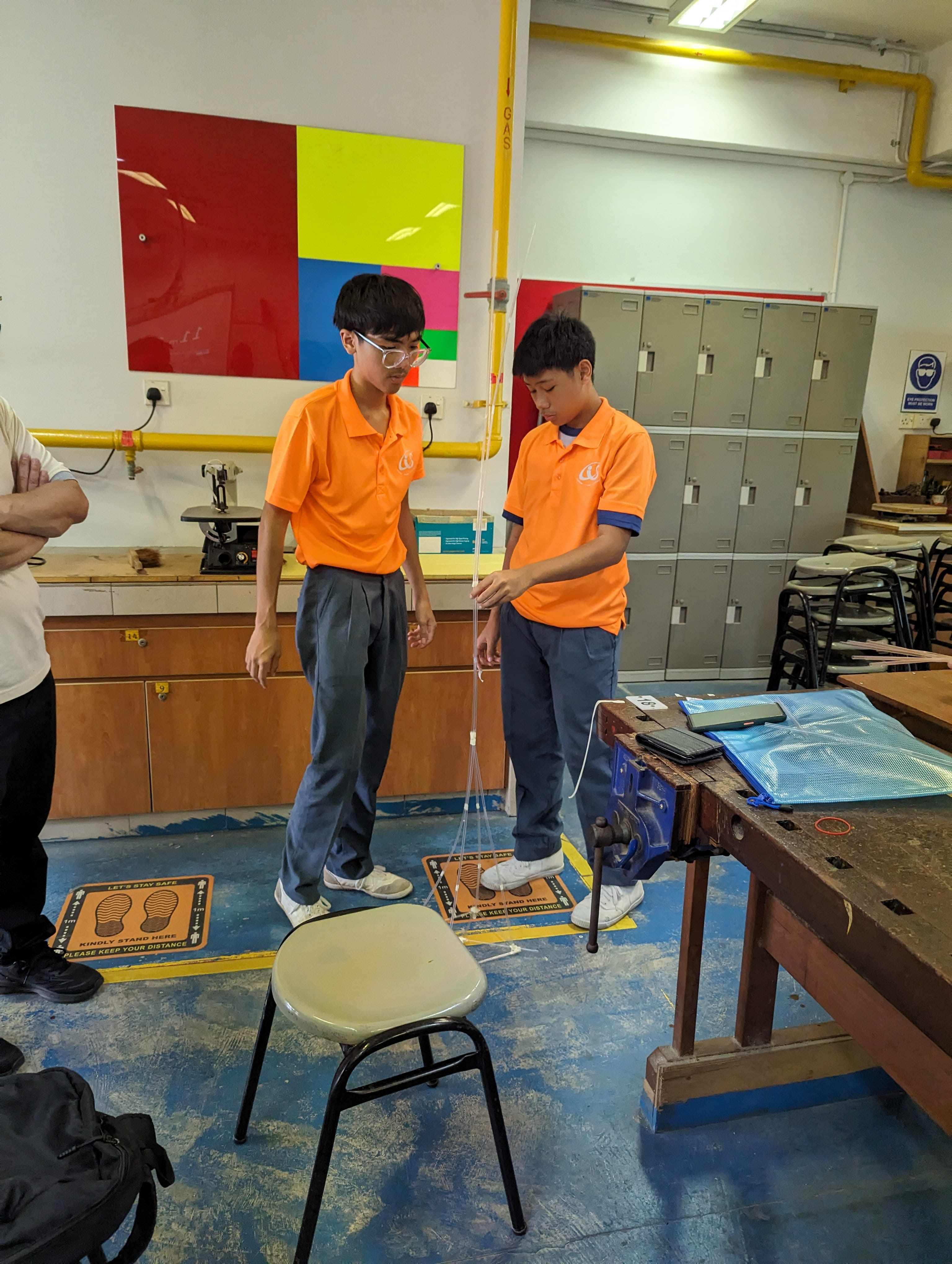 Two students in orange uniforms work with a string and stool in a school workshop with lockers and workbenches.