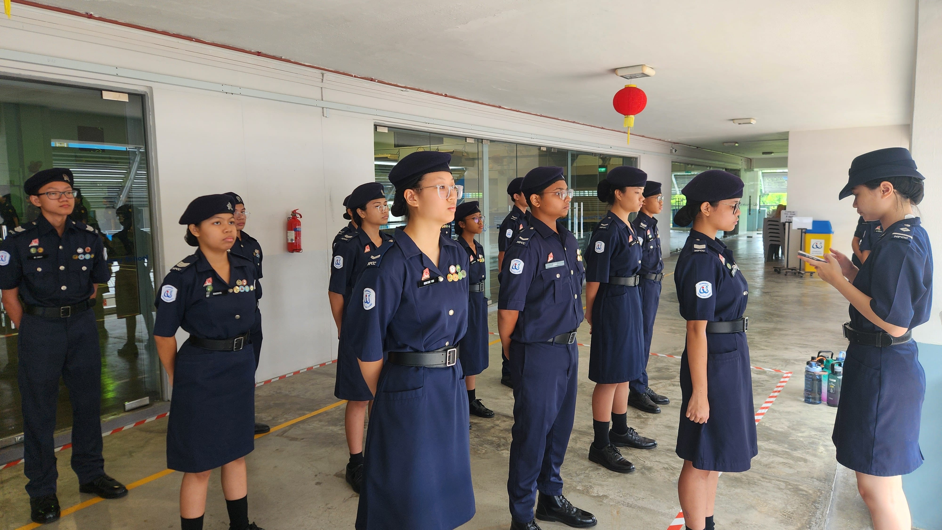 Young cadets in navy blue uniforms and berets stand in formation indoors while one reads from a clipboard.
