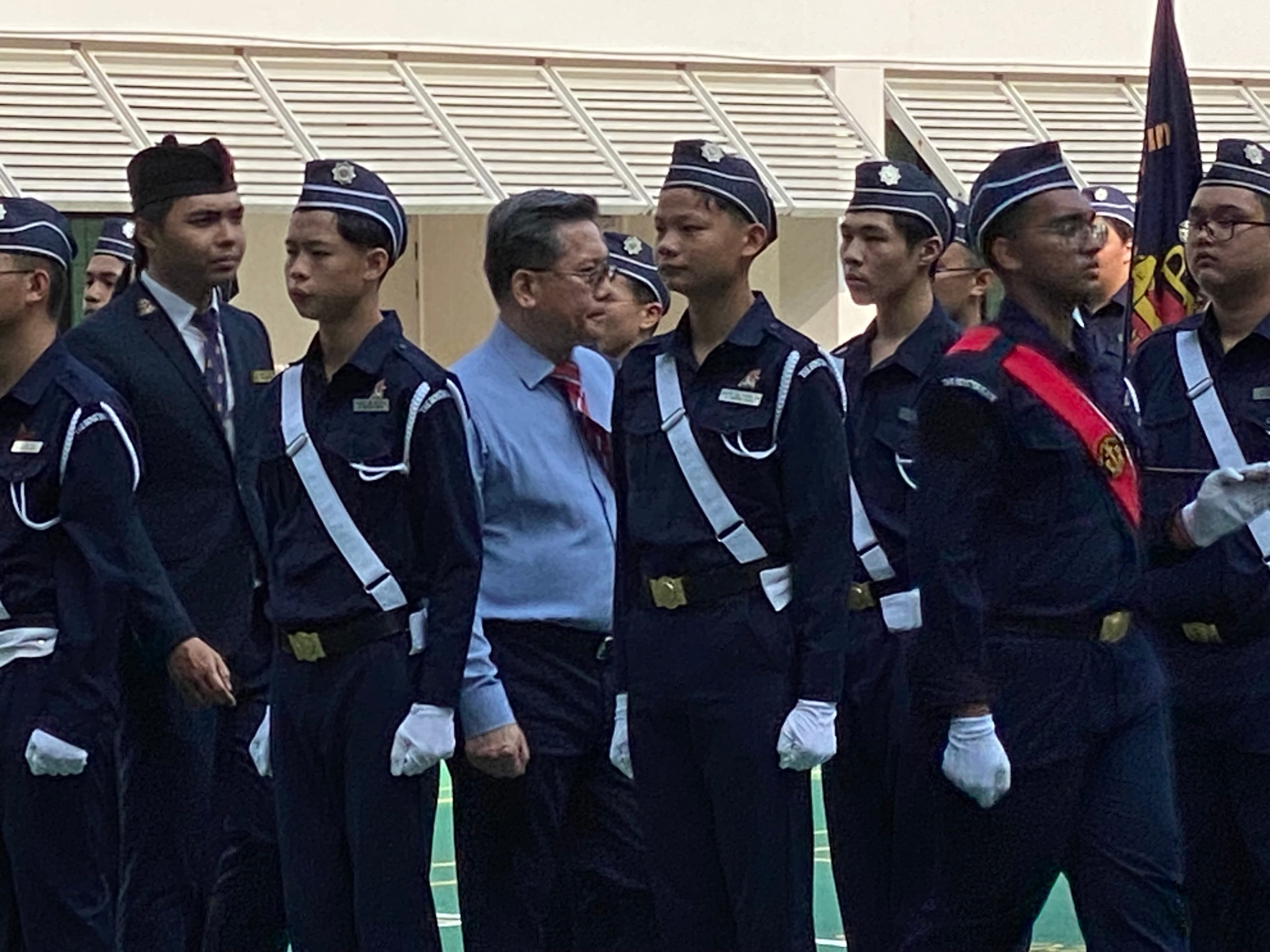 A civilian in a blue shirt stands among uniformed cadets in dark navy uniforms with white gloves and a flag bearer.