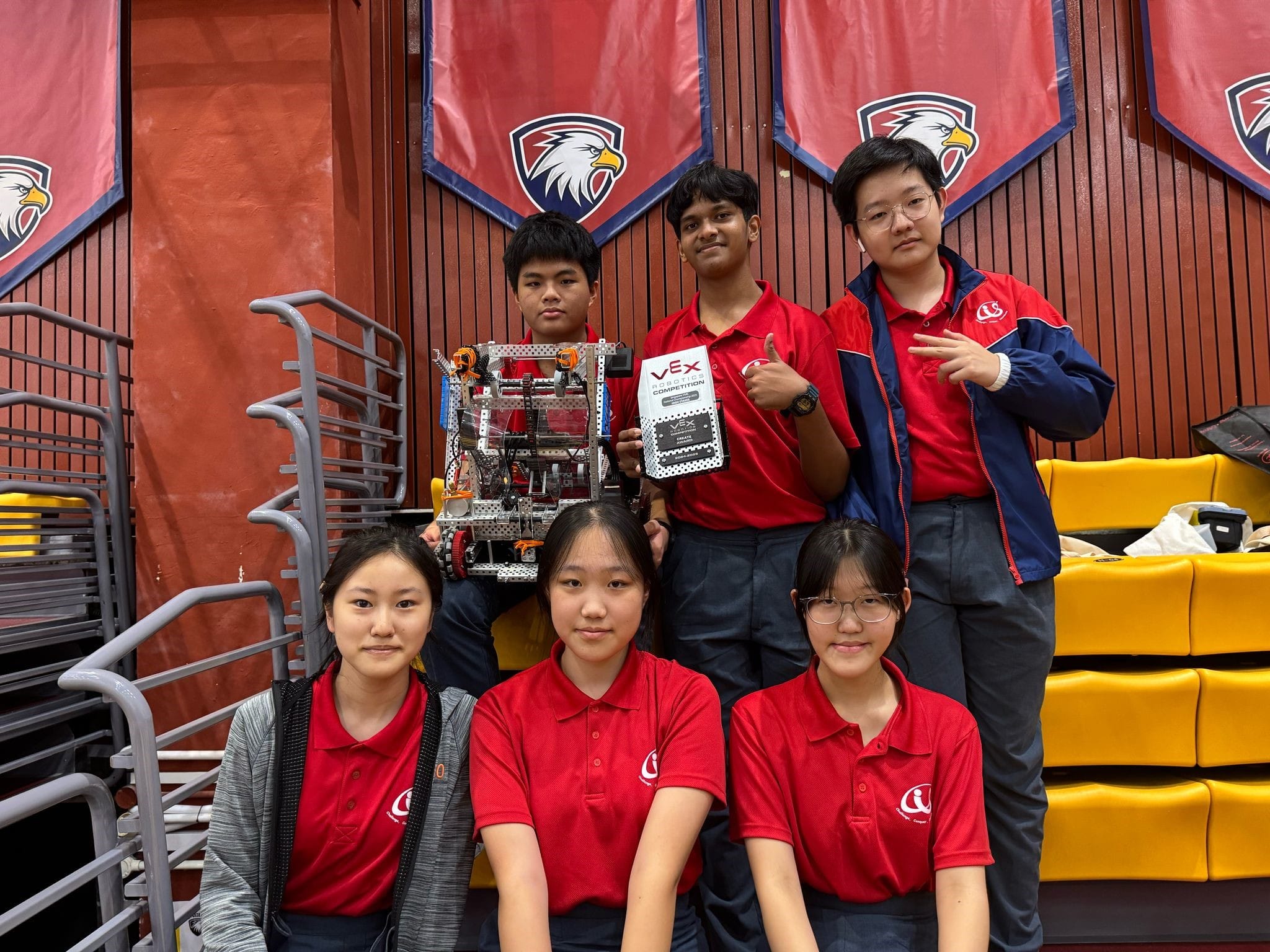 Six students in red uniforms pose with a VEX Robotics Competition robot and award in a gym with eagle banners.