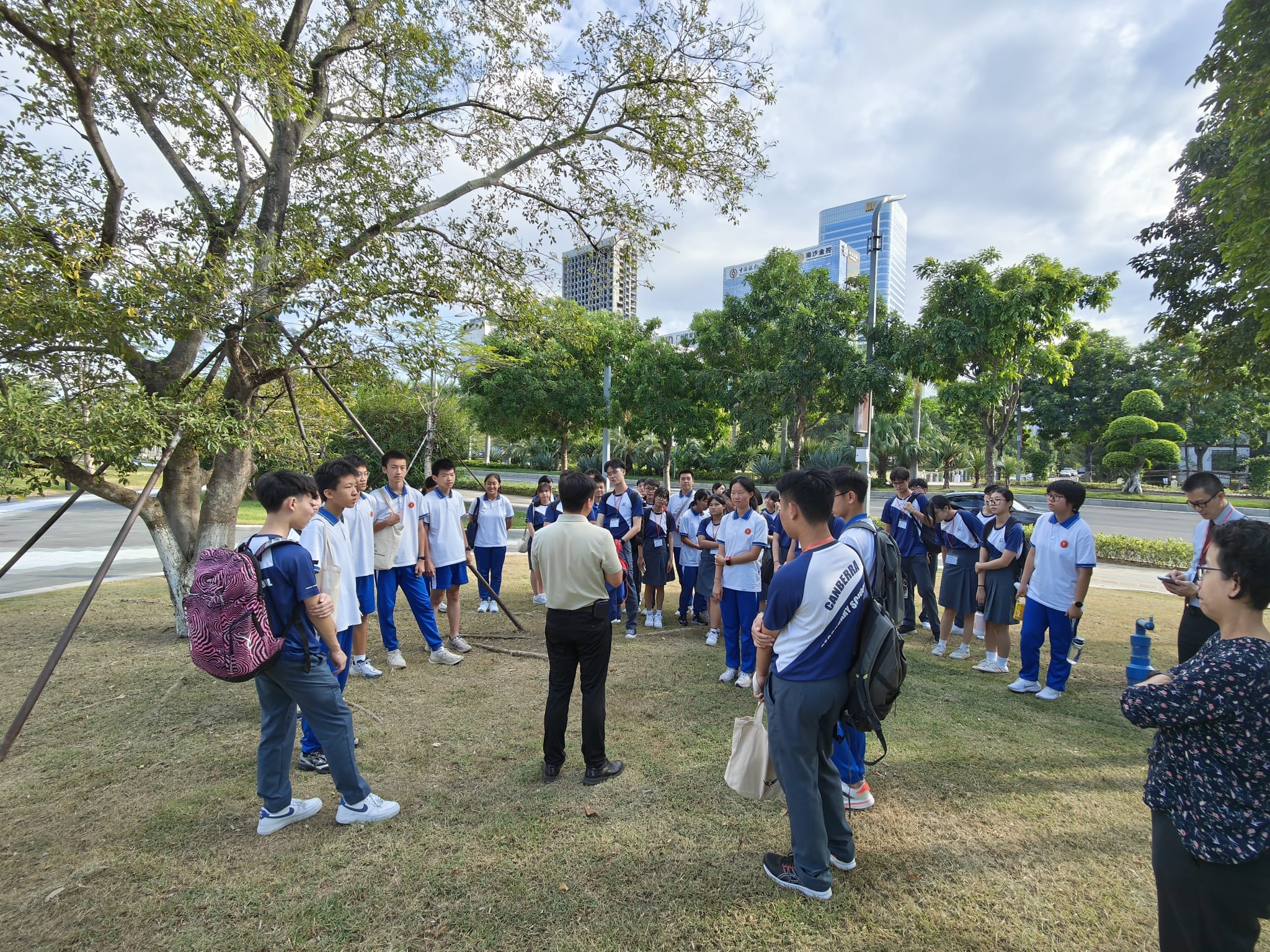 A group of students in white and blue uniforms gather around an adult guide outdoors in a park with city buildings behind.