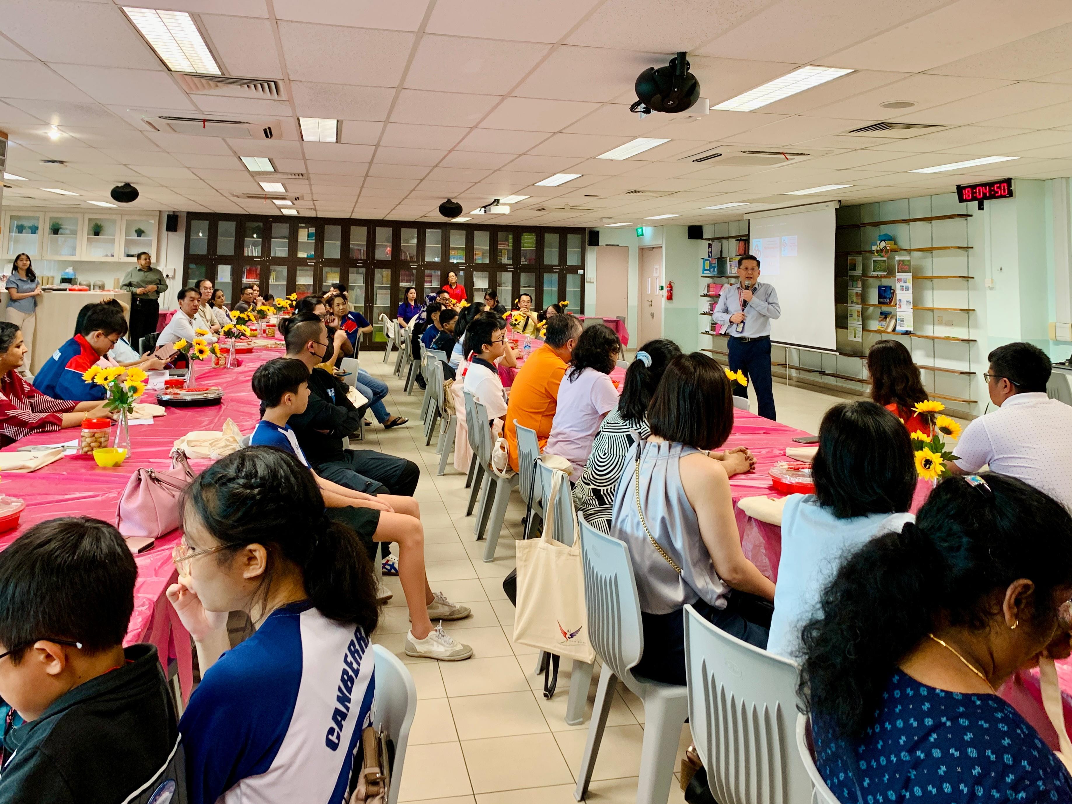 A man speaks at a microphone to a large group seated at pink-covered tables decorated with sunflowers indoors.