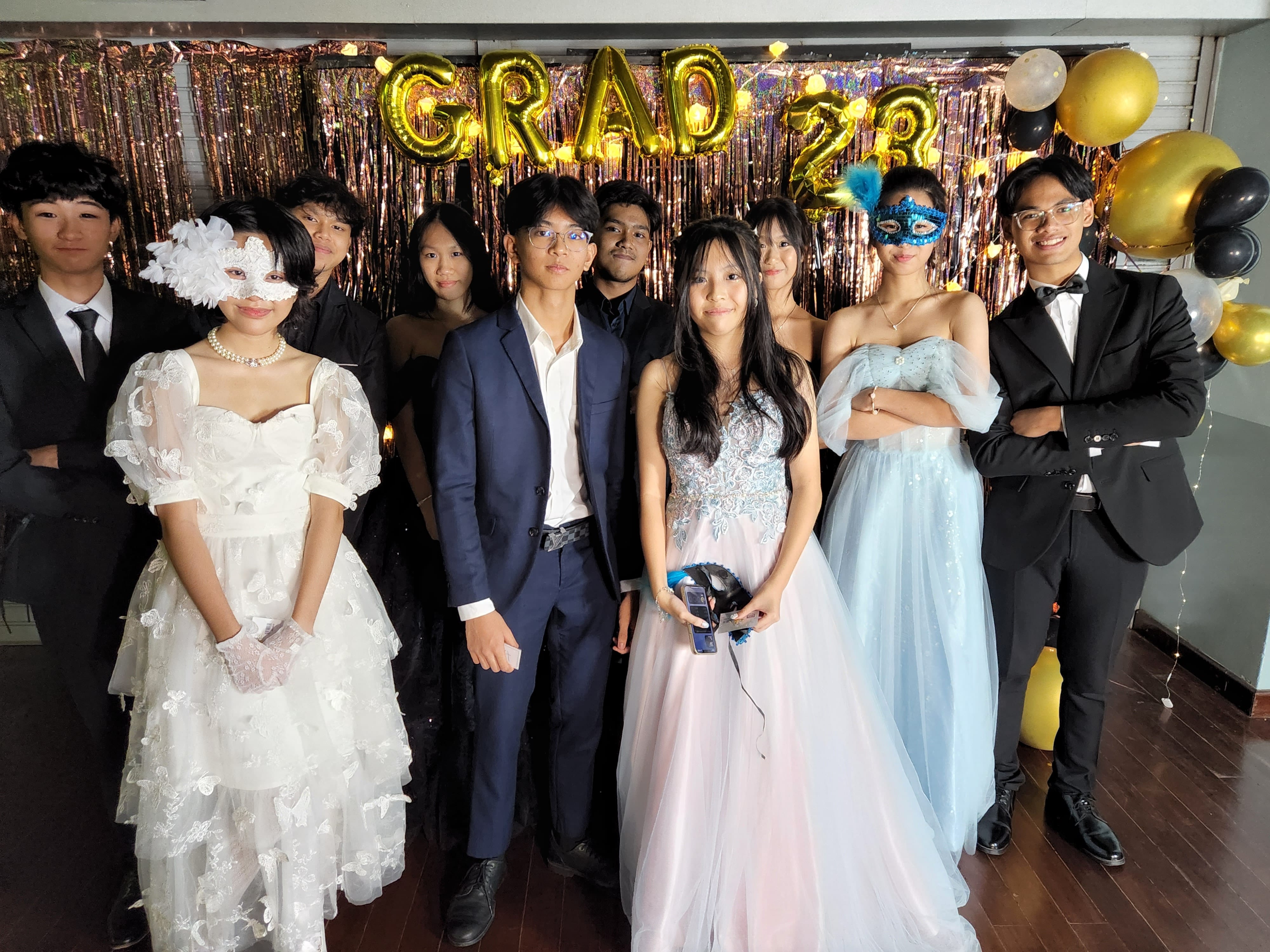 A group of formally dressed teens pose at a graduation party with gold GRAD balloons and a shiny backdrop.