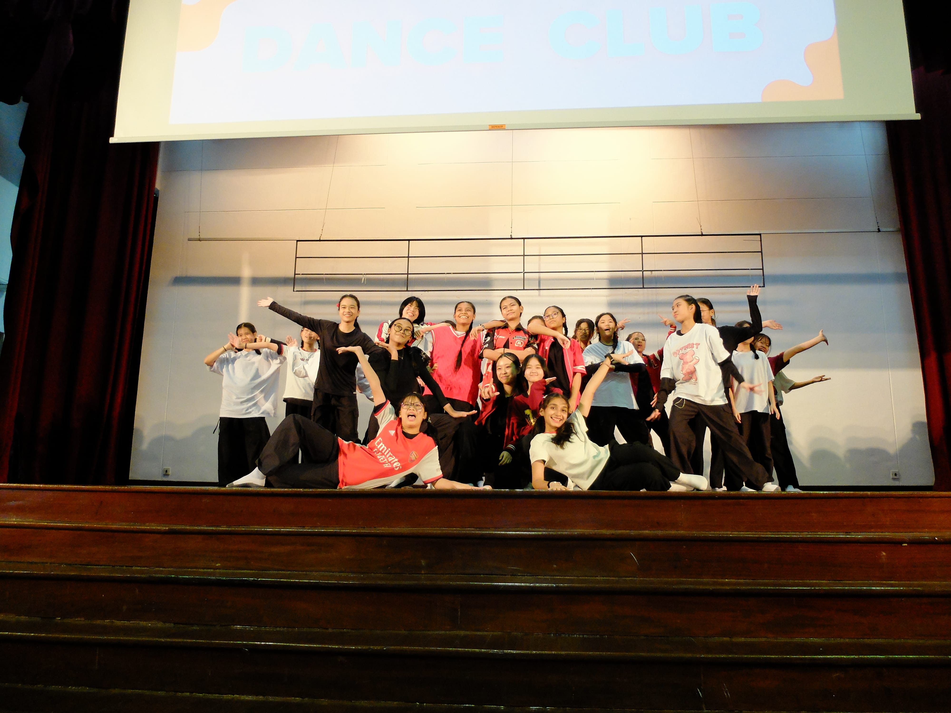 A group of young students pose on a stage under a projected screen reading Dance Club.