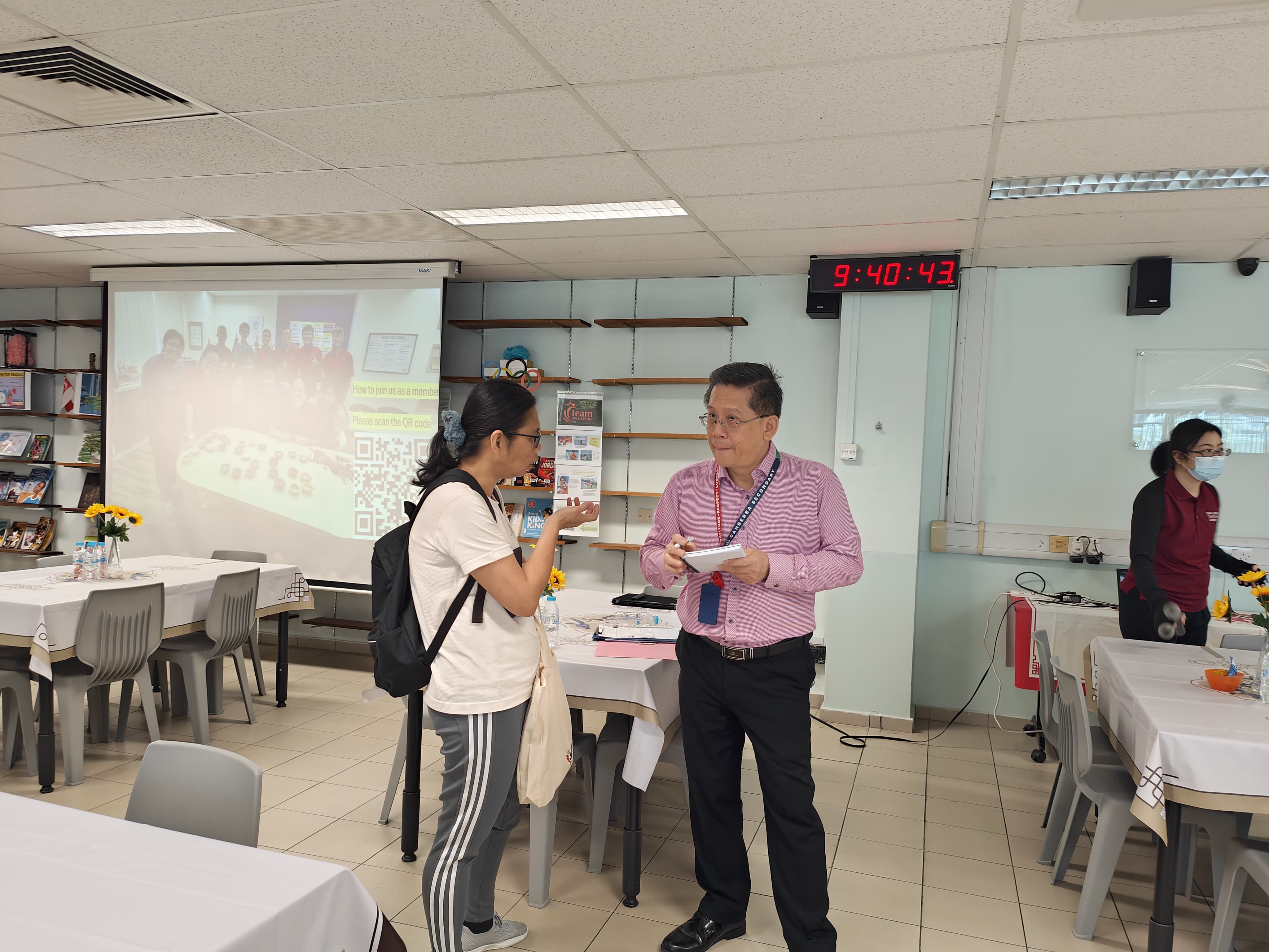 Two people talk in a meeting room with a projected QR code screen and a digital clock showing 9:40:43.