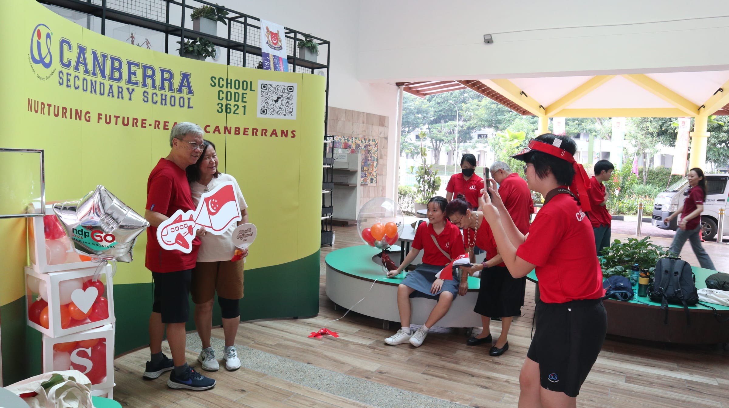 Two people pose with Singapore-themed props at a Canberra Secondary School event while a student photographs them.