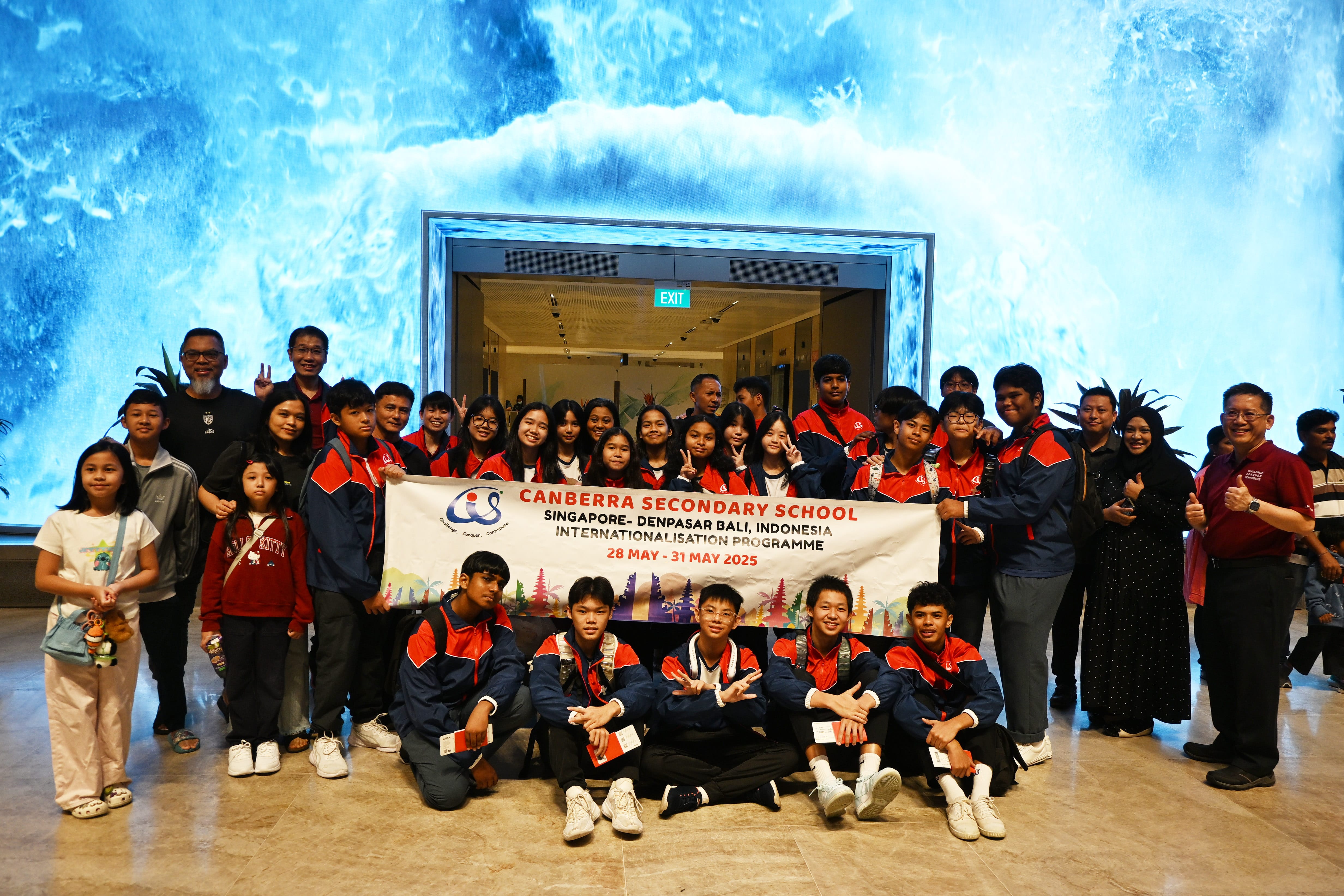 A large group of students in navy and red uniforms pose with a Canberra Secondary School Singapore-Bali programme banner.