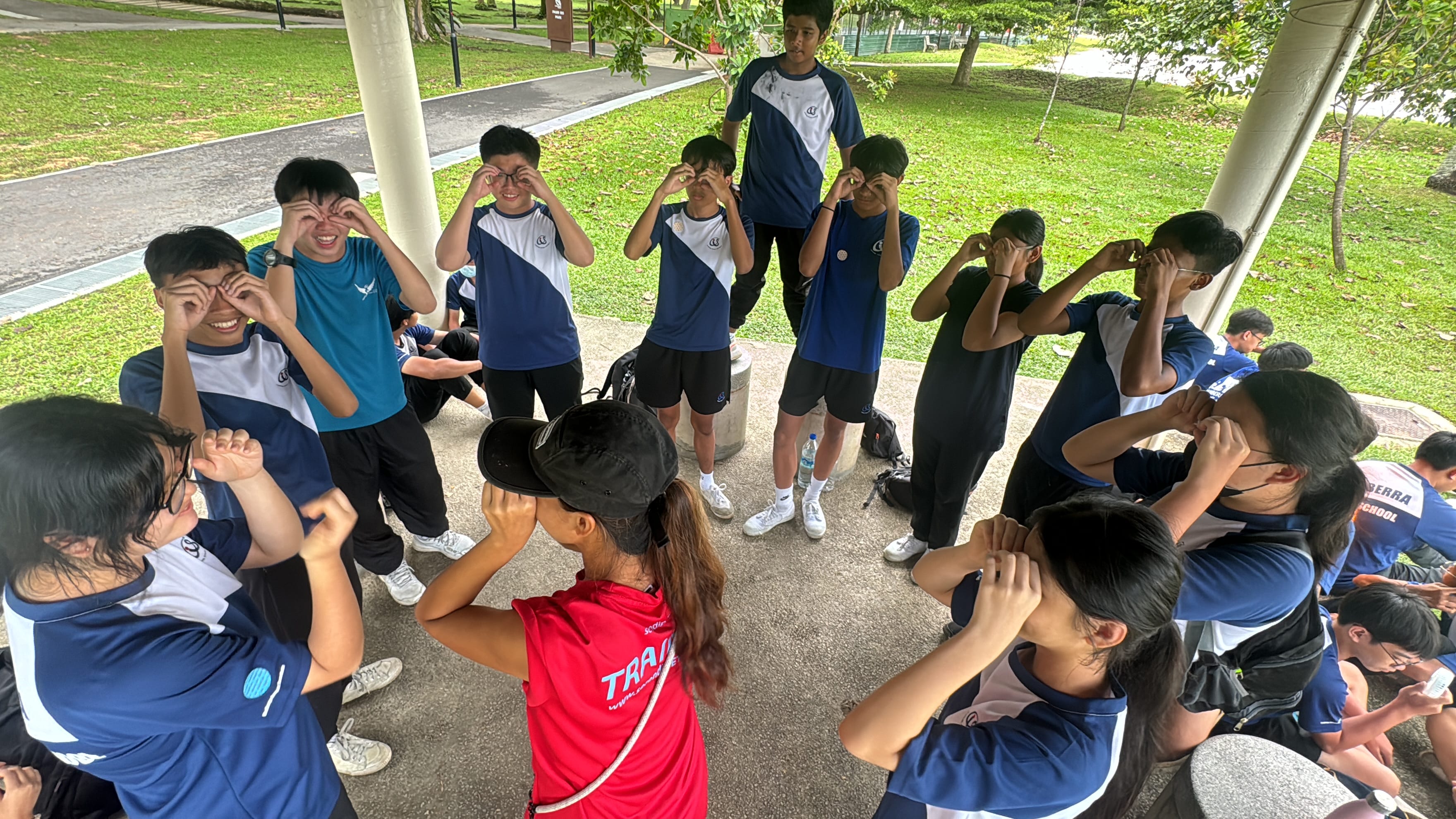 A group of students in blue uniforms stand in a circle outdoors, making binocular gestures with their hands around their eyes.