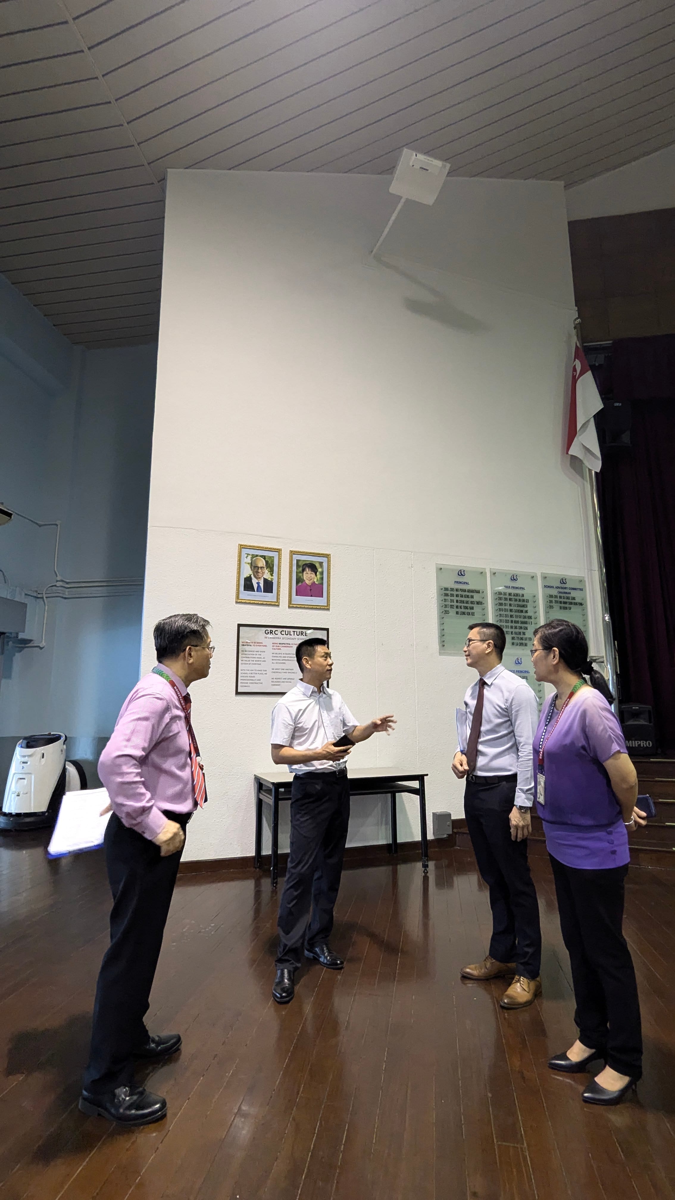 Four people in business attire talk on a wooden floor in a hall with plaques and portraits on the wall.