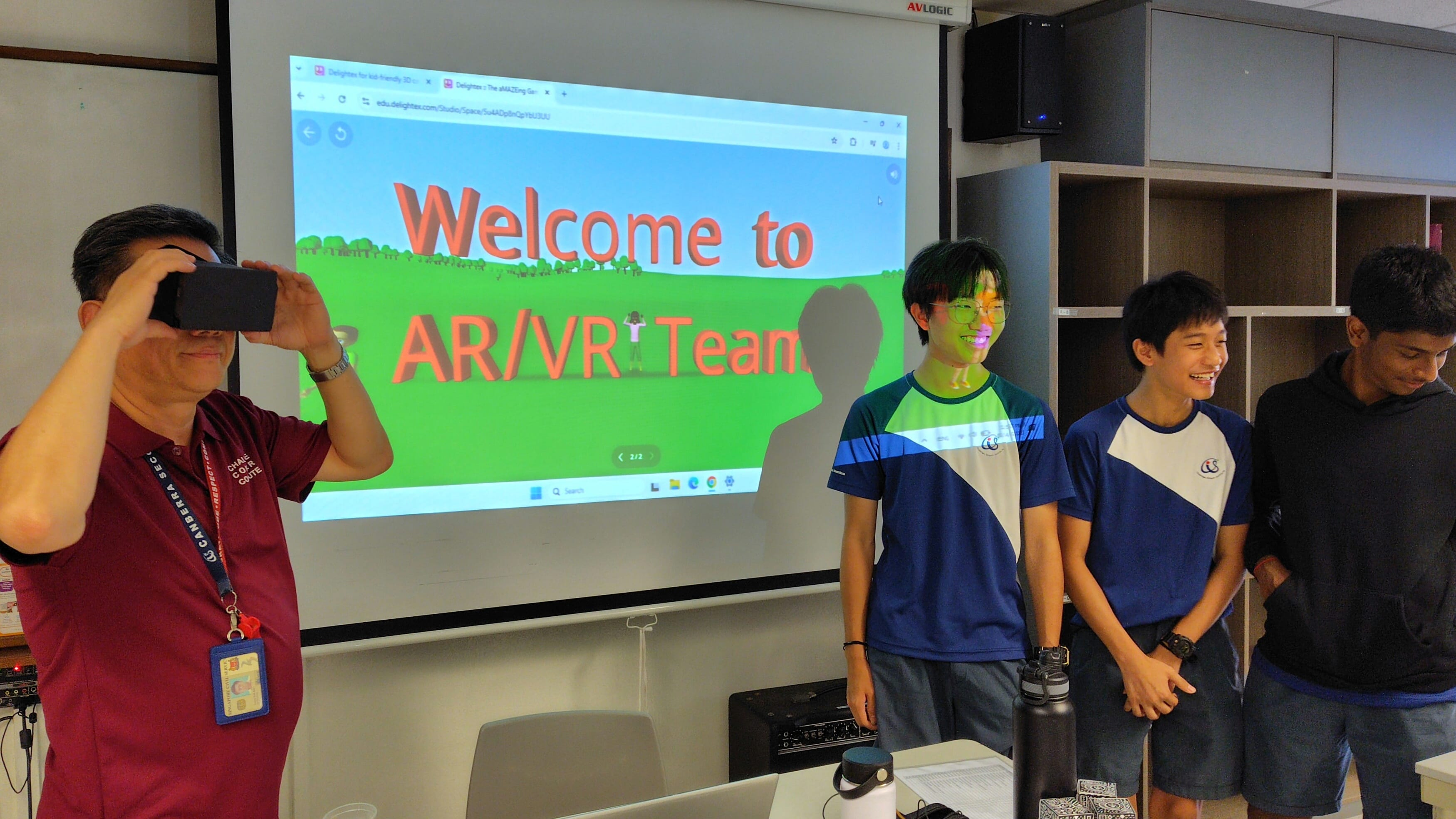 An adult holds a VR viewer to their face while three students stand smiling before a projected Welcome to AR/VR Team slide.