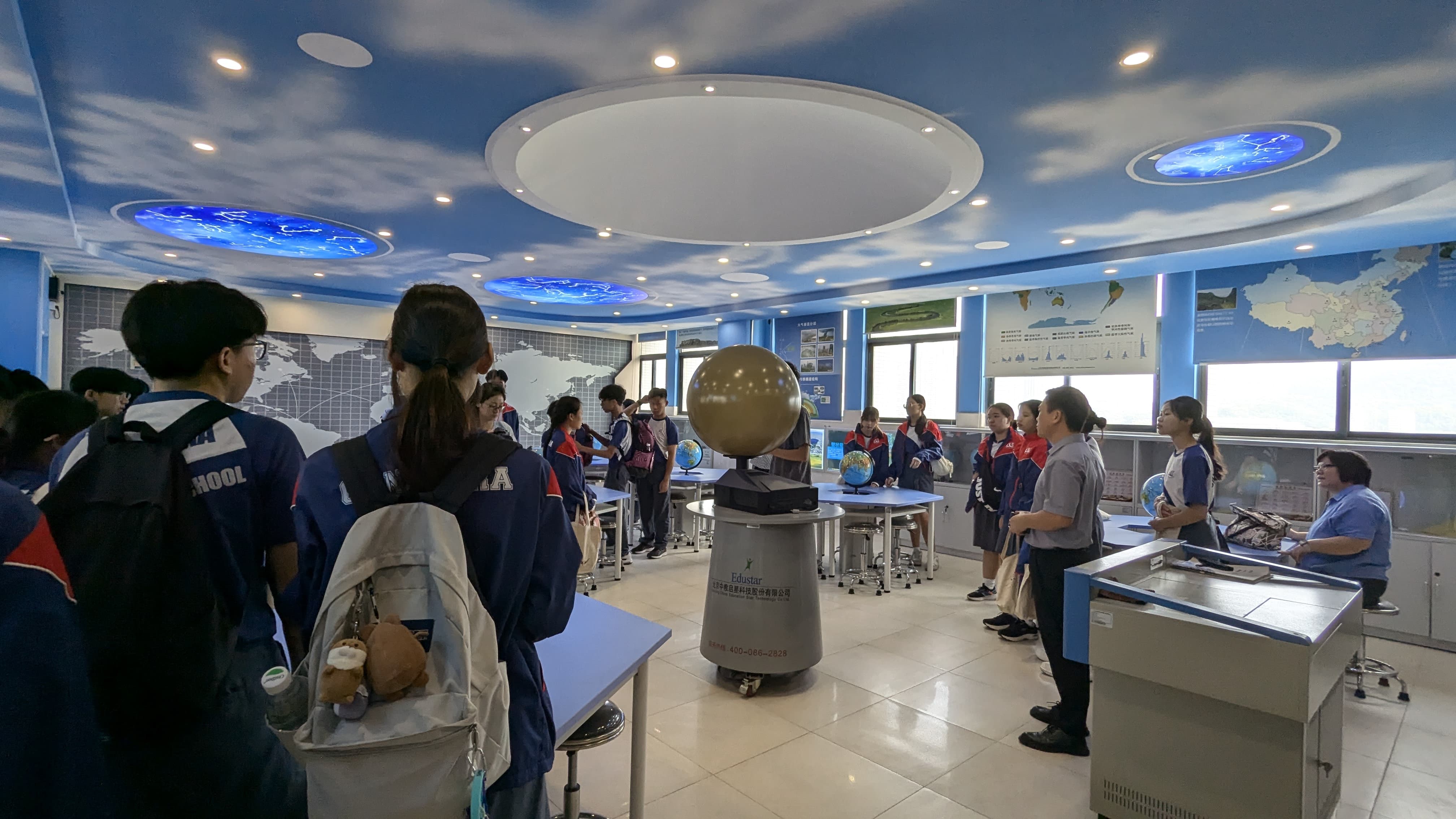 Students in uniforms explore a geography classroom with globe displays, map walls, and a blue sky-themed ceiling.