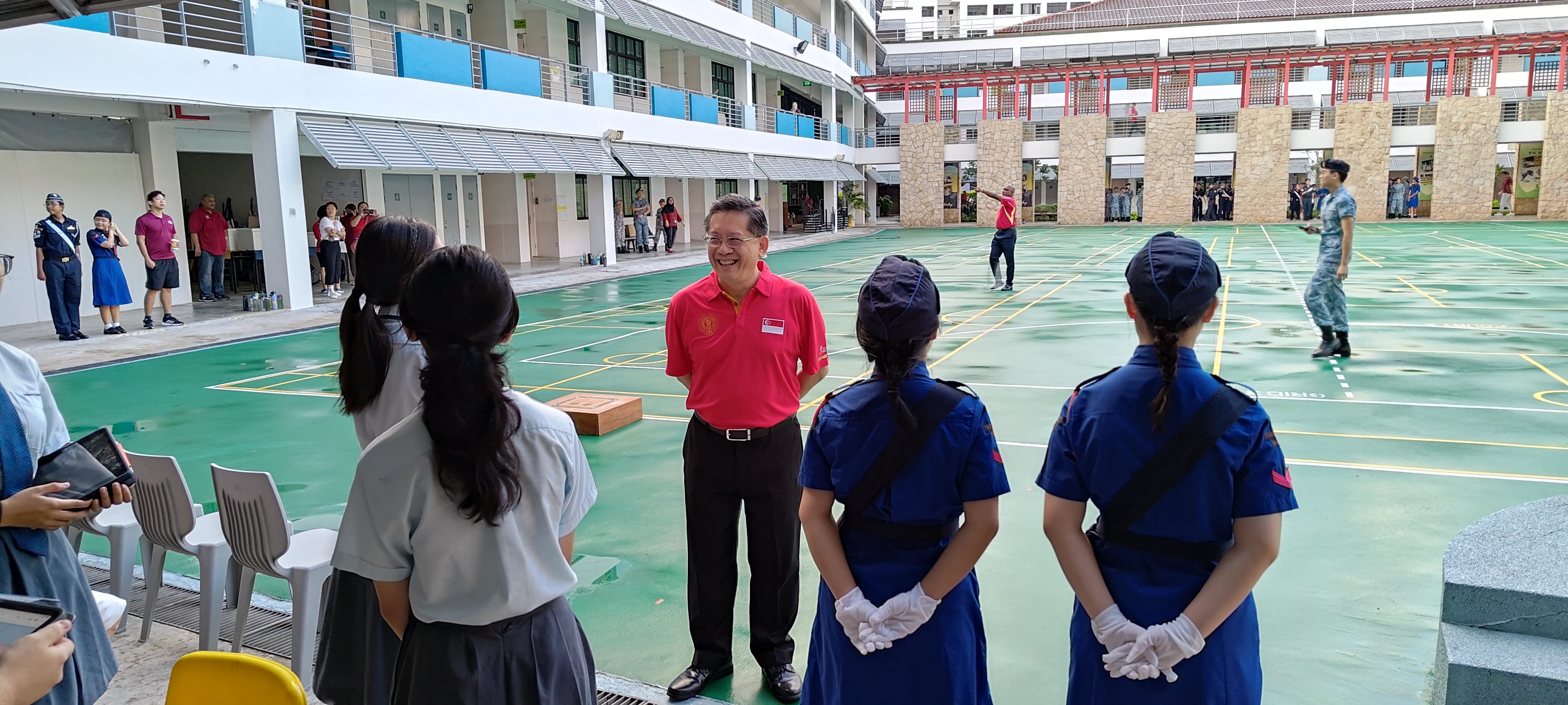 A smiling man in a red polo speaks with students in blue uniforms and white gloves on a school courtyard.